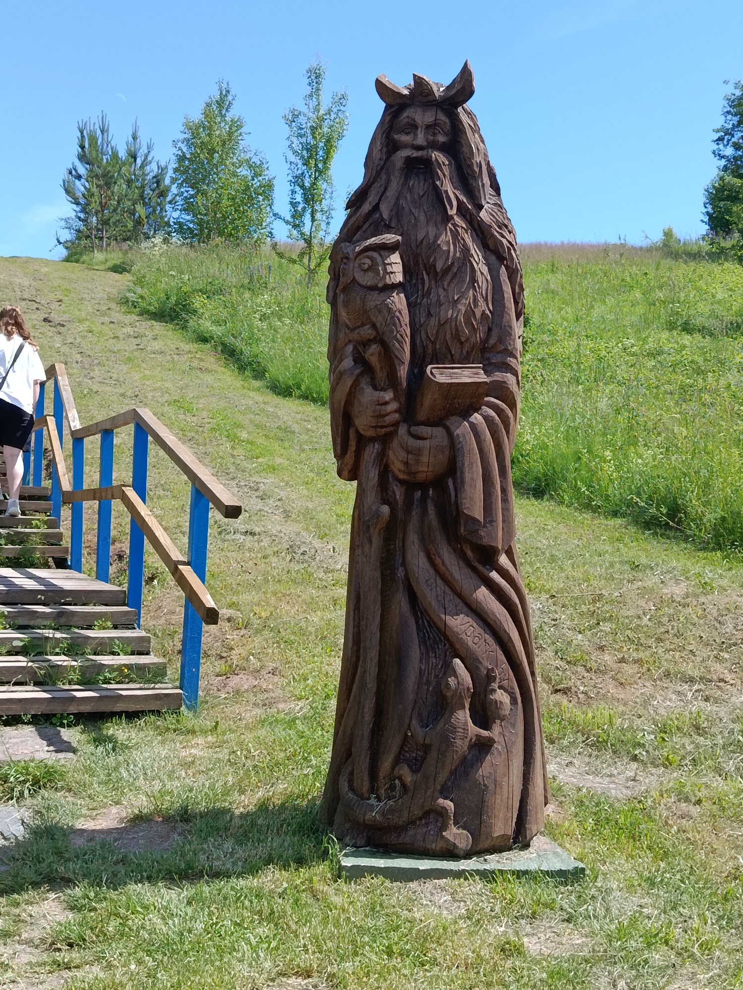 A wooden sculpture of a wizard holding an owl and a book stands in front of a grassy hillside under a clear blue sky. The wizard is carved with a long beard, a crown-like headdress, and flowing robes. At the base of the sculpture, smaller carvings of animals are visible. To the left of the sculpture, a wooden staircase with blue railings ascends the hillside, with a person in a white shirt and black shorts walking up the stairs. The environment is natural, with green grass and scattered trees on the hill. The location is identified as Plyuski village, Belarus.