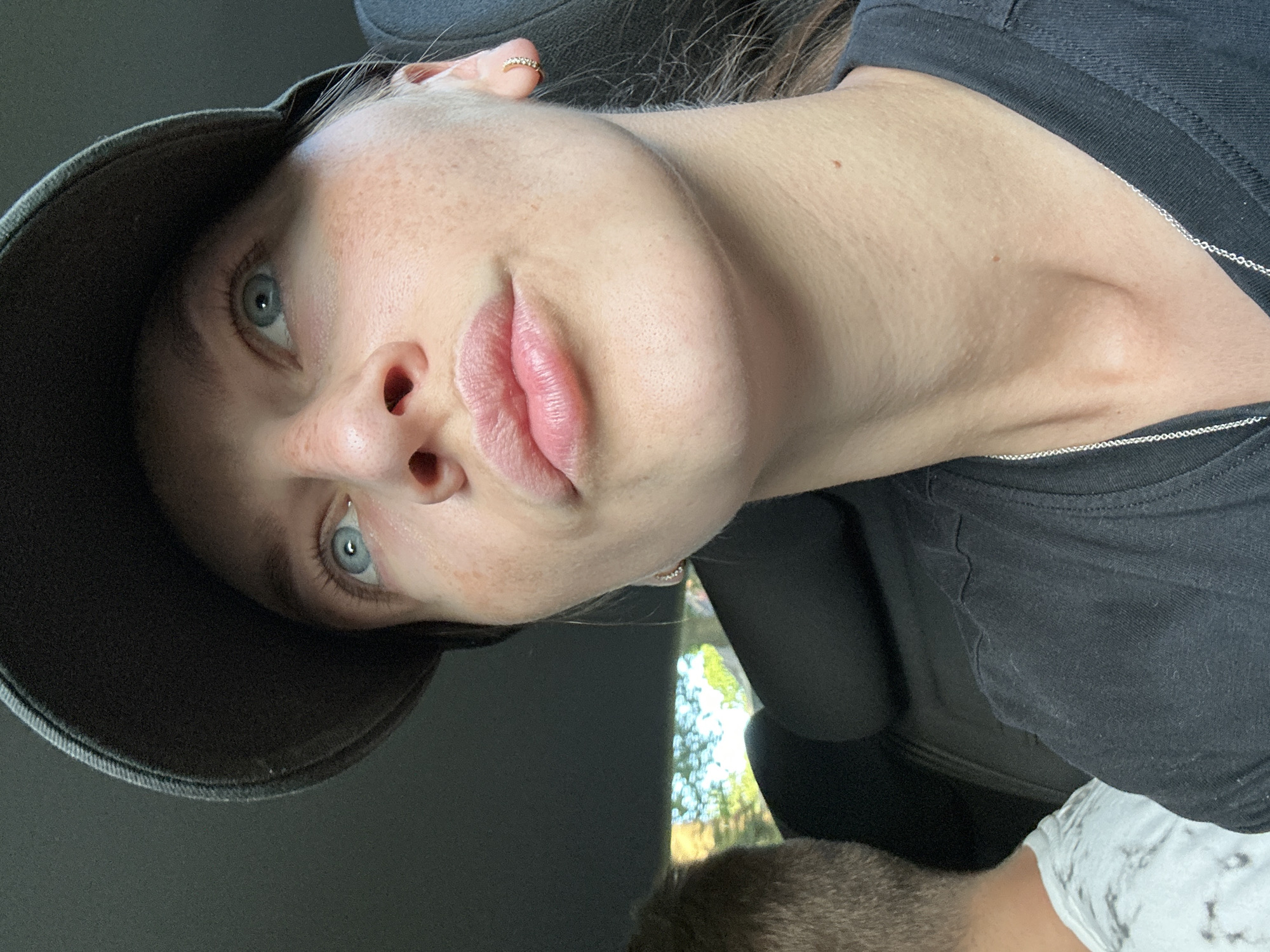 A selfie-style photograph shows a fair-skinned individual with freckles and light blue eyes, wearing a dark-colored baseball cap and a black t-shirt. A silver necklace is visible. The person appears to be in a car. The location and city of Bargstedt, Germany cannot be confirmed from the image alone.