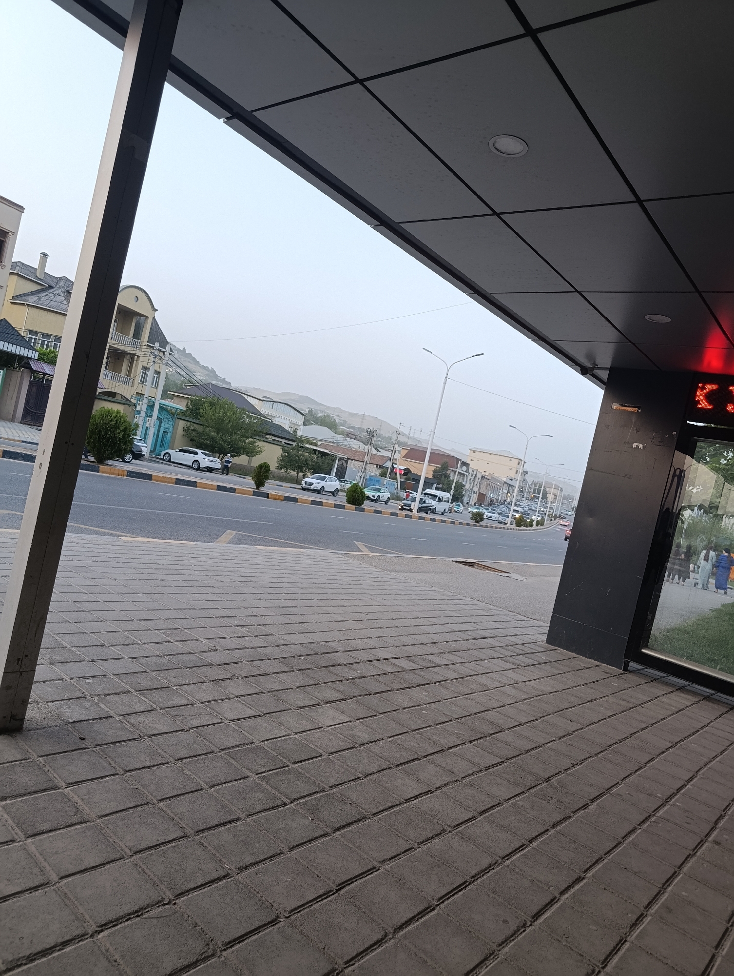 The image depicts a street scene in Dushanbe, Tajikistan, likely during the daytime, with an overcast or hazy sky suggesting it might be early morning or late afternoon, or due to atmospheric conditions. The perspective is from a sidewalk or covered walkway, looking out towards a road and buildings.
In the foreground, a textured gray tiled pavement leads towards a modern building with a dark facade. Part of this building, likely a storefront or entrance, features a reflective glass window and a glowing red display sign with some visible text, though it is largely obscured and difficult to read. A thick, light-colored pillar is also prominent in the foreground, part of the building's structure.
The street in the middle ground is busy with traffic, including several cars, a white van, and a small bus. People are visible walking on the sidewalk across the street. The architecture of the buildings lining the street varies, with some appearing residential with balconies and others more commercial. Trees and streetlights are also present. In the background, hills or mountains can be seen under the hazy sky. The overall impression is a typical urban environment.