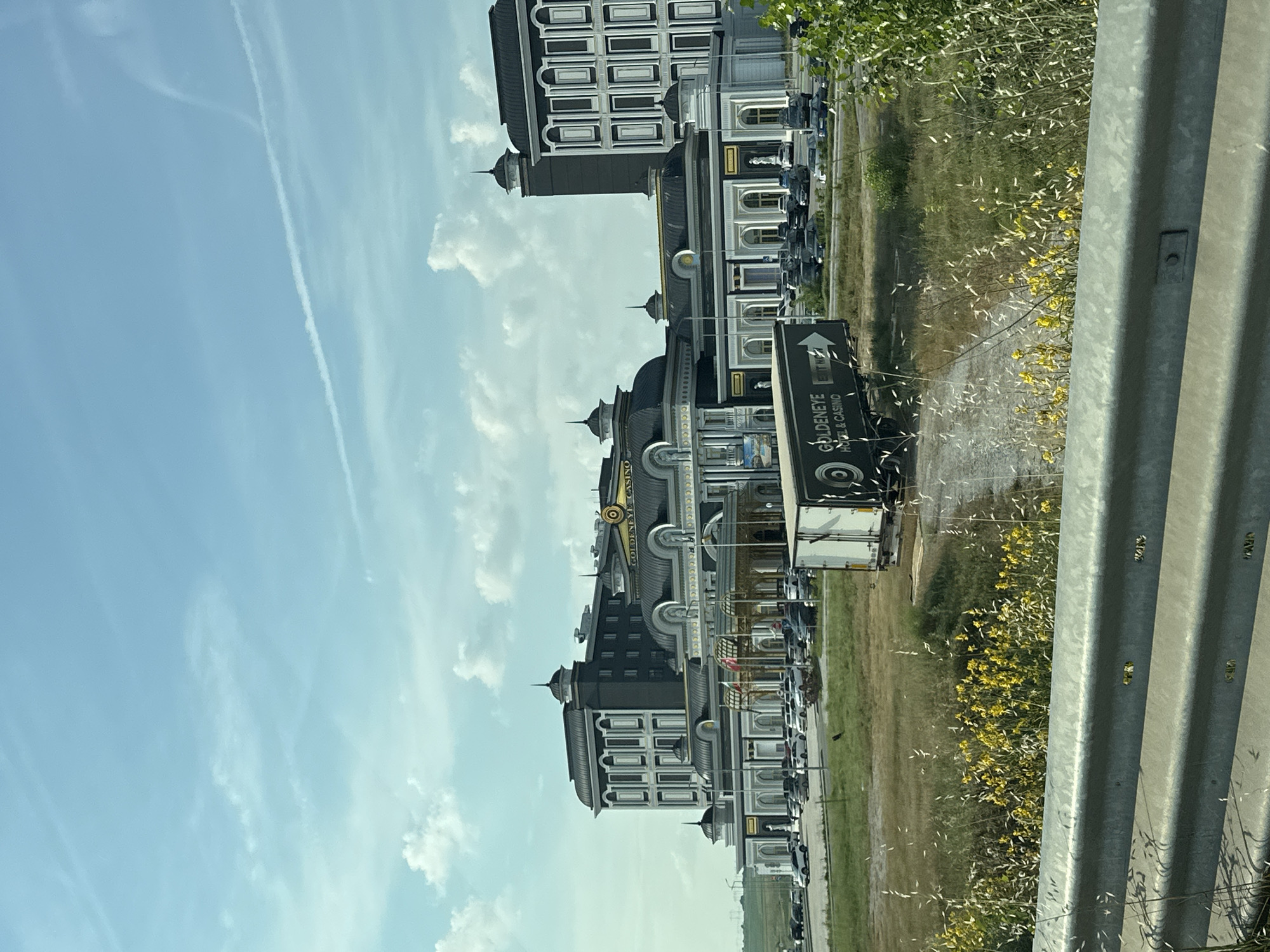 The image captures the Goldeneye Hotel & Casino in Kapitan Andreevo, Bulgaria, from a roadside perspective on a clear day with scattered clouds. In the foreground, a grey metal guardrail runs horizontally across the bottom, behind which is an unkempt grassy field with prominent yellow flowering plants and tall dry grasses. A dirt path cuts through this vegetation, leading towards the casino complex. A dark-colored semi-trailer truck, bearing the "GOLDENEYE HOTEL & CASINO" logo on its side, is positioned on this path in the mid-ground.
The casino complex is a large, multi-story structure exhibiting ornate, classical architectural styling. It features dark grey domed roofs with gold accents and light-colored facades. The main central section of the building prominently displays "GOLDENEYE CASINO" signage near its roofline. To the left and right, taller, rectangular wings with multiple windows and decorative elements flank the central structure. A grand covered entrance with columns is visible in the central section, with what appear to be flags flying nearby. A large paved parking lot in front of the casino is occupied by numerous parked cars. In the far left background, beyond the casino, power lines and pylons are faintly visible against the horizon. The sky is bright blue with scattered white cumulus clouds and several faint white contrails from aircraft. No individuals are clearly discernible in the image.