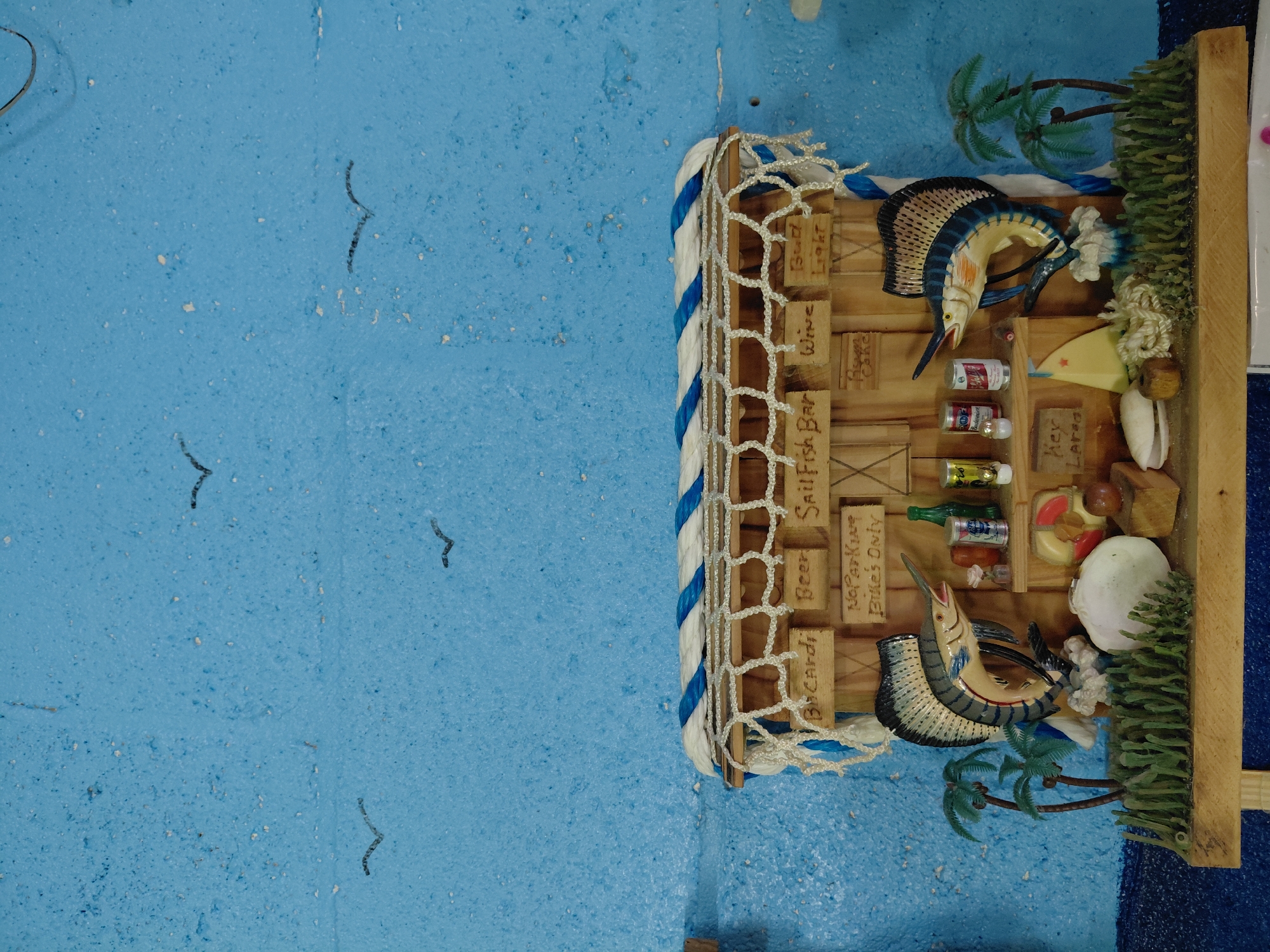 The image depicts a miniature, three-dimensional diorama of a coastal bar setting. Key features include wooden signage indicating offerings such as "Beer," "Wine," "Sailfish Bar," "Bud Light," and "Bacardi." The bar is adorned with miniature bottles and cans, along with nautical decorative elements such as marlin fish figurines, seashells, and artificial palm trees and foliage. The structure of the bar appears to be made of wood, wrapped with nautical rope and netting. The entire scene is placed against a textured, light blue surface, possibly representing the ocean or sky.
The location of Cutler Bay, United States of America is provided solely as context for the image and is not visually discernible within the image itself.