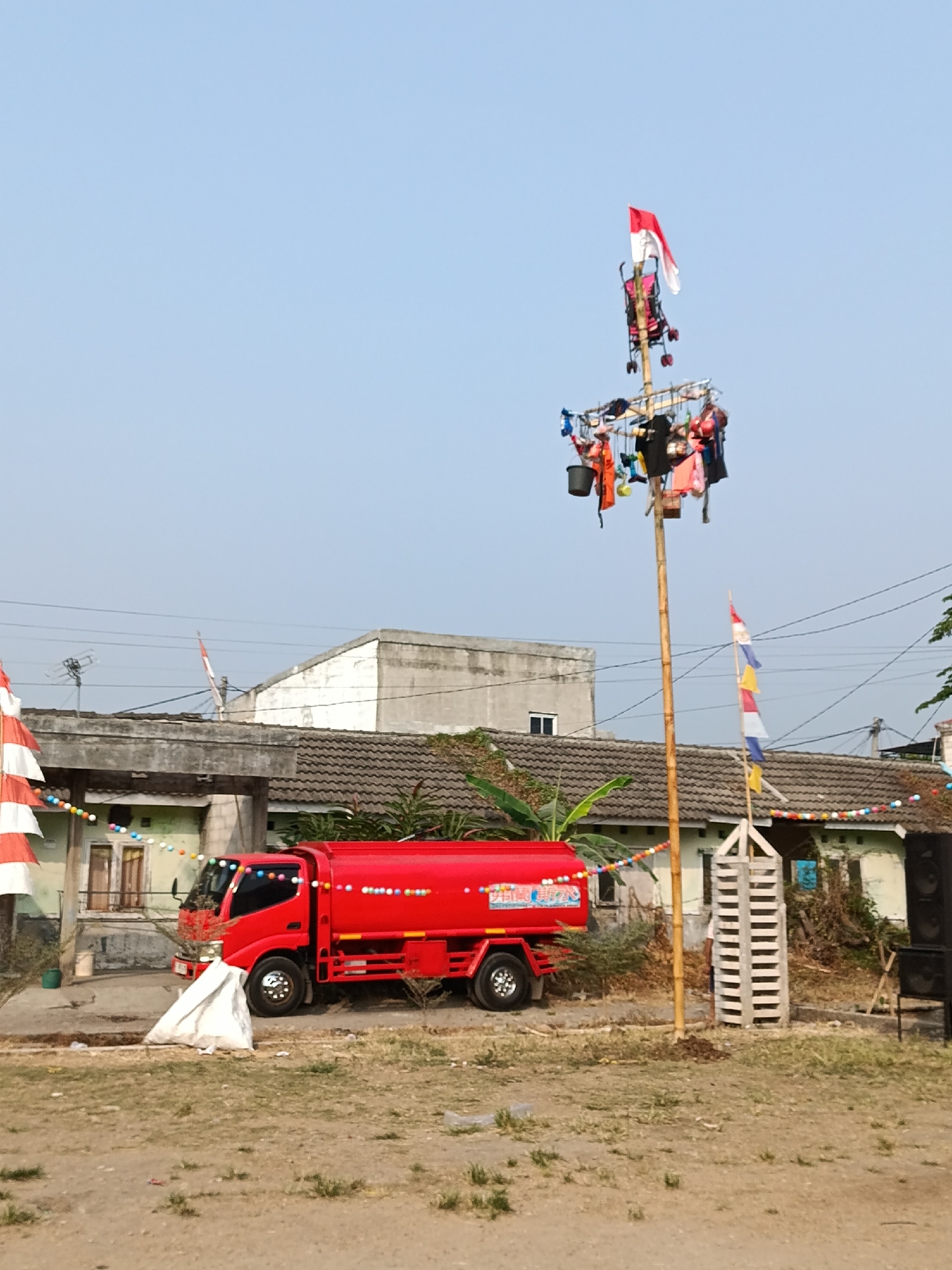 The image shows a street scene in Cariu, Indonesia, likely during a celebration. In the foreground, a bright red truck with a water tank is parked. To the right of the truck, a tall bamboo pole stands, adorned with multiple Indonesian flags of varying sizes and colors, as well as various objects hanging from it, including buckets and clothing items. A smaller, white, wooden structure with horizontal slats is placed at the base of the bamboo pole. In the background, there are buildings with tiled roofs and a clear blue sky. Decorative strings of colorful lights are strung across the scene, between the buildings and the pole.