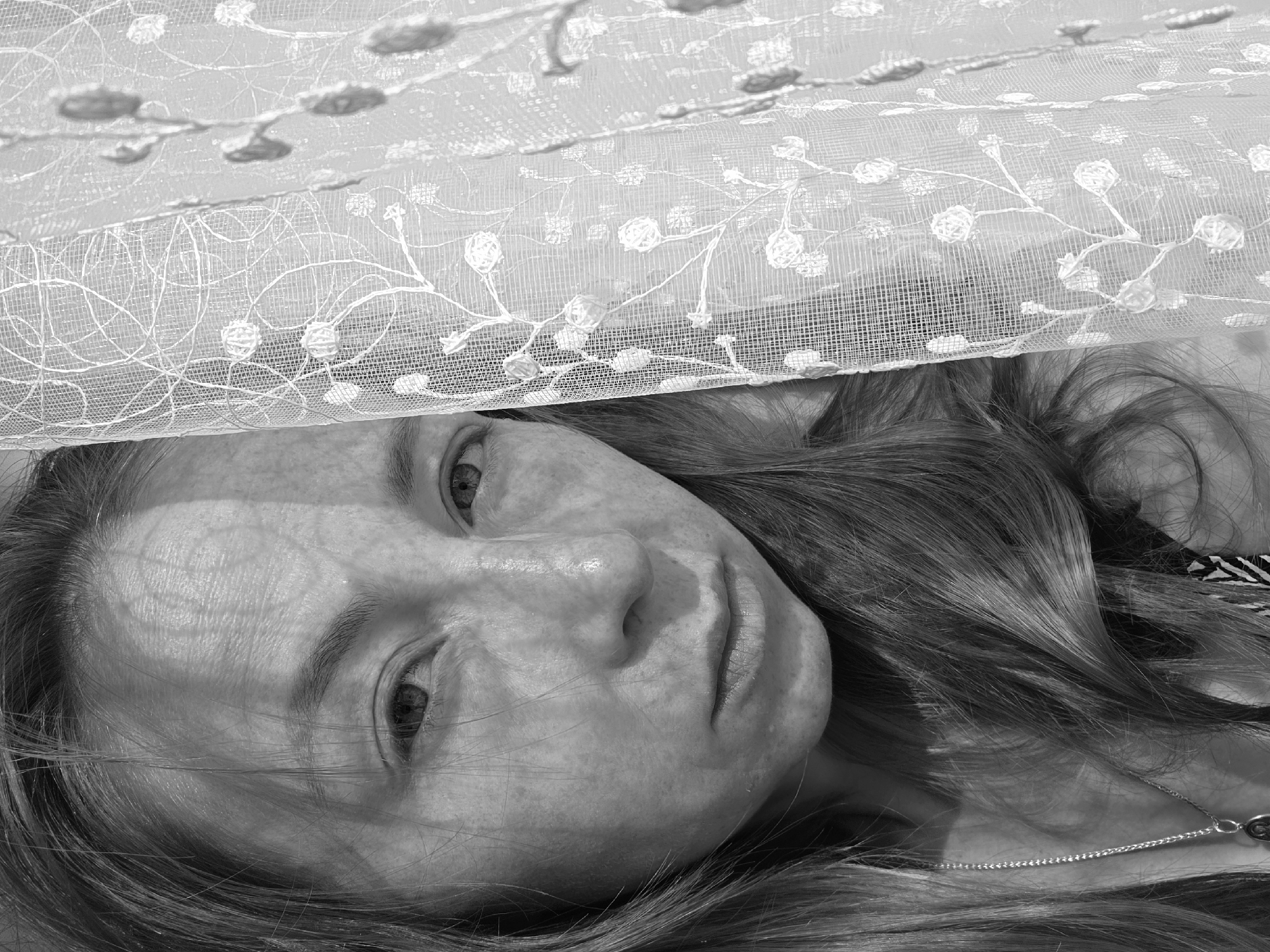 A black and white close-up image features a woman with light skin and long hair, lying down beneath a textured fabric. The fabric, possibly a curtain or netting, has a pattern of floral designs. The woman's face is partially obscured by the fabric. She has light eyes and a necklace is visible around her neck. The location is Unknown, Unknown.