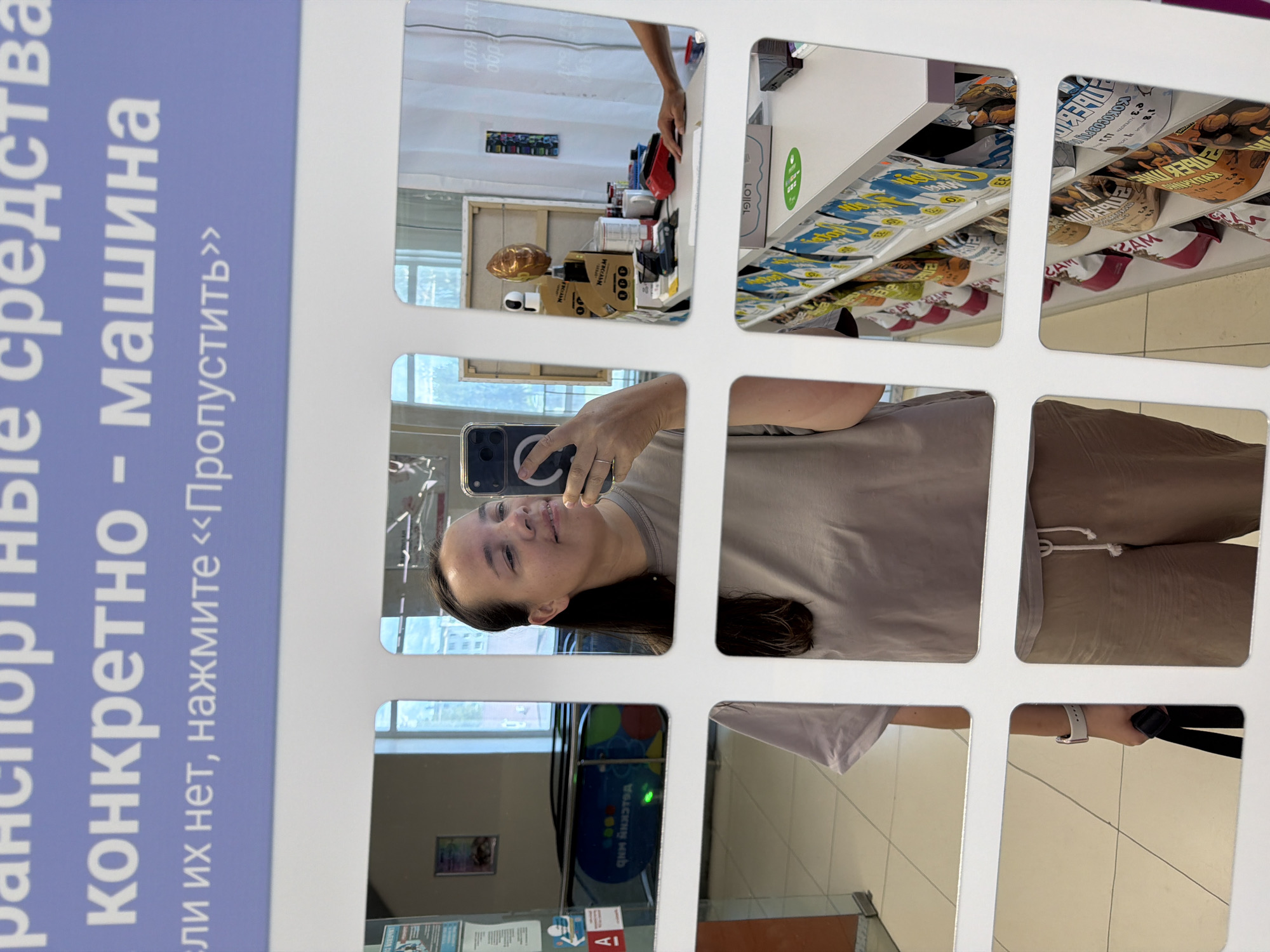 The image shows a selfie taken by a woman in a retail environment that is likely a supermarket in Chaikovsky, Russia, based on the Cyrillic text on the signage. The woman is holding a smartphone in her right hand, using it to capture her reflection in a display of multiple mirrors. She is wearing a light brown t-shirt and beige shorts.
In the background, reflected in the mirrors, are various elements of the store. One mirror shows an escalator, a poster with the Russian word "АНКЕТА" (ANKETA), and a red and white "A" symbol. Another reflection reveals shelves stocked with packaged goods, possibly snacks or food items, with Cyrillic labeling visible. There's also a glimpse of a counter with various items like boxes, cups, and what appears to be a stapler or tape dispenser. In the lower portion of the image, the woman's legs and feet are visible, along with a black bag she is holding in her left hand. The overall impression is a casual snapshot taken within a shopping area.