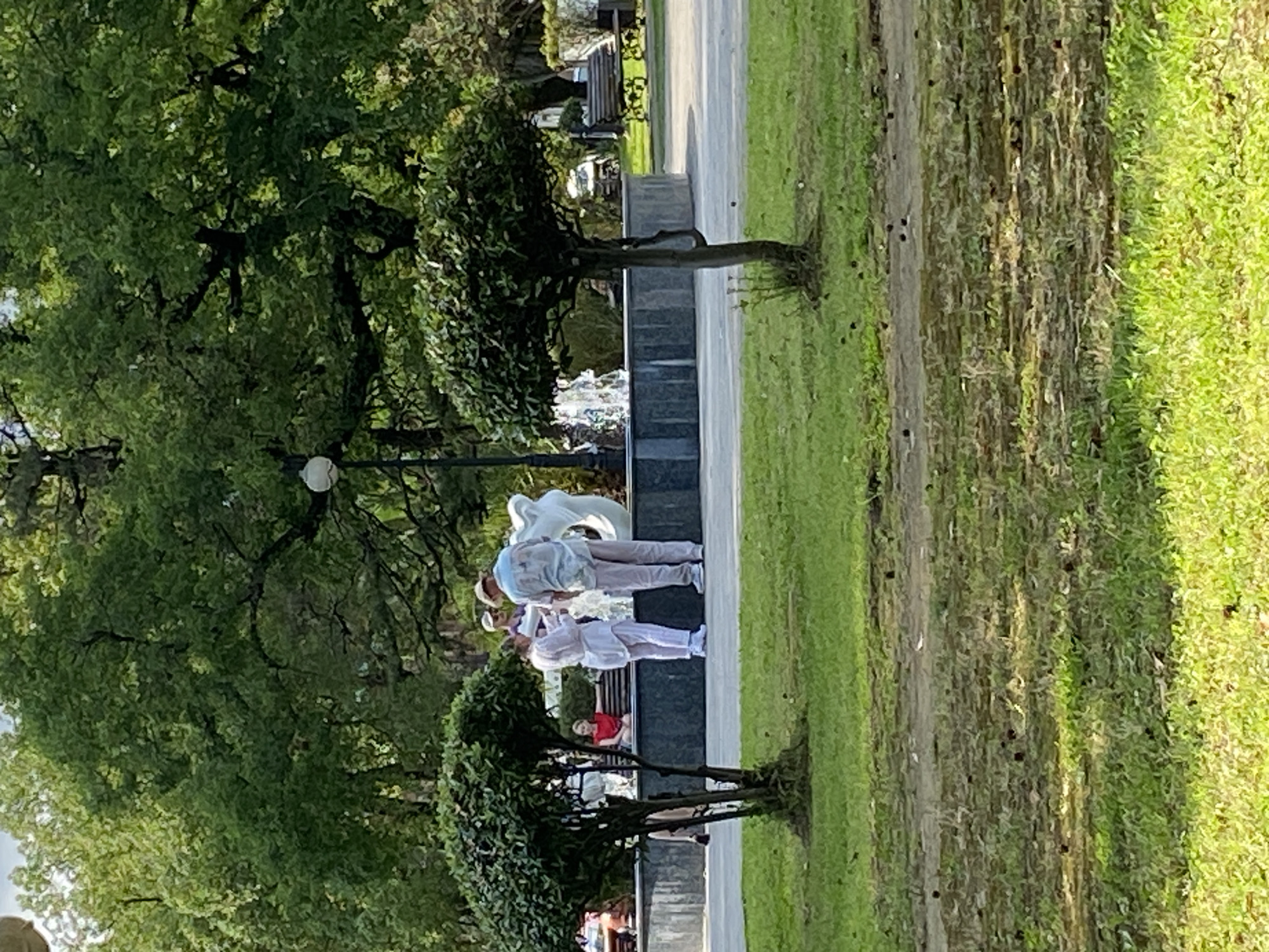 In Sokhumi, Abkhazia, at the fountain area near the seaside boulevard, two individuals are engaged in conversation near a white abstract sculpture. The man is wearing a light-colored patterned shirt and light-colored pants, with a hat. The woman is wearing a white outfit. In the background, a fountain sprays water. To the left, a woman in a red top sits on a bench. The foreground features a manicured lawn with small trees and scattered debris. The environment suggests a public park with lush greenery and a pedestrian walkway.