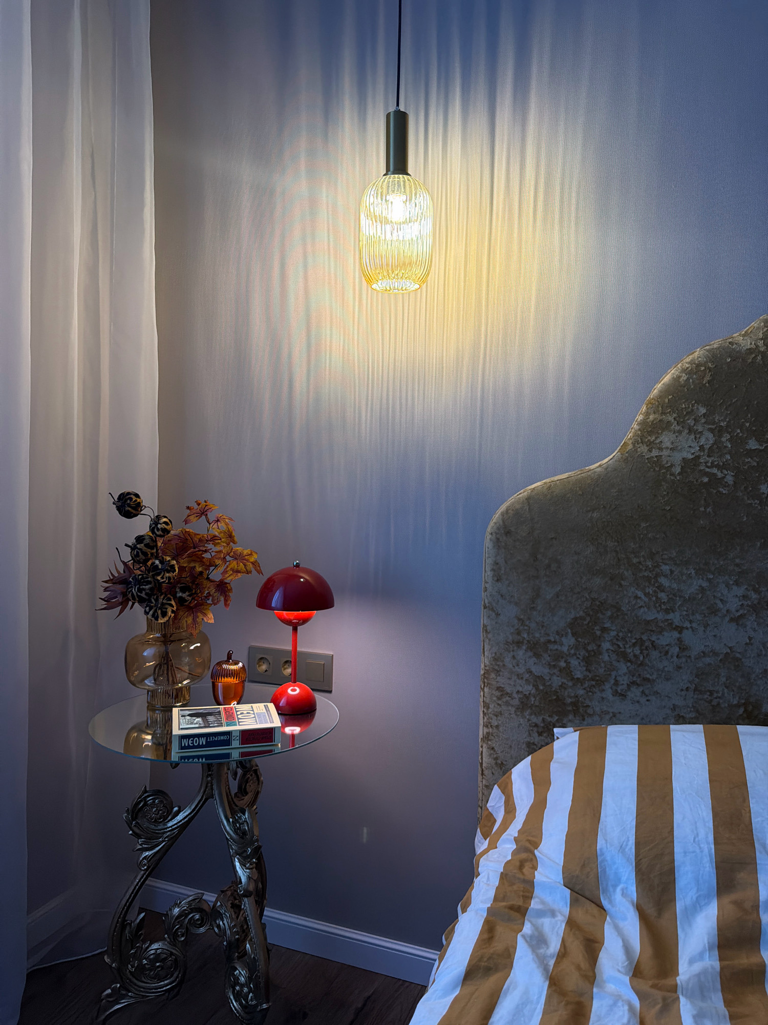 A bedside scene in a room, possibly in Orsha, Belarus, features a small round glass table with ornate golden legs. On the table, a glass vase holds a dried floral arrangement. To its right, a small amber-colored decorative acorn sits next to a red mushroom-shaped table lamp. A book, titled "MEOW", lies flat on the table. A white sheer curtain is visible on the left, and a bed with a textured, gold-toned headboard and striped bedding (white and golden brown) is on the right. The room is lit by a single hanging light fixture above, casting elongated, striped shadows on the wall. Electrical outlets and a switch are visible on the wall behind the lamp. The floor appears to be dark wood. There are no people present in the image.
