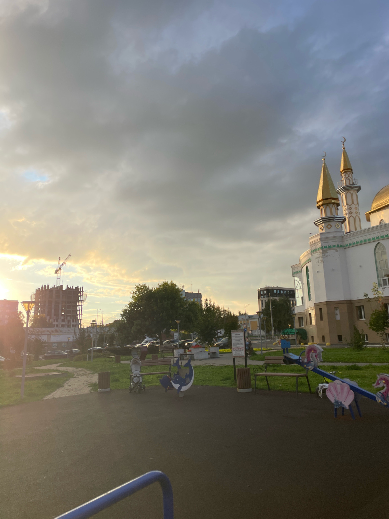 The image depicts a playground and a modern mosque in Izhevsk, Russia, under a cloudy sky with sunlight breaking through. In the foreground, a black asphalt playground surface is visible, with a blue railing in the lower left corner. A seesaw with pink and blue seats, shaped like seashells and horses, is set up on the grass. Nearby, a stroller and a bin are present. Behind the playground, a paved path winds through a grassy area with trees. In the middle ground, a construction site with a crane and an unfinished building stands to the left, while cars are parked along a road. The prominent mosque, with its white facade, green trim, and golden domes and minarets topped with crescents, dominates the right side of the frame. The sky is filled with dramatic, grey clouds, interspersed with areas of golden sunlight, suggesting either sunrise or sunset.