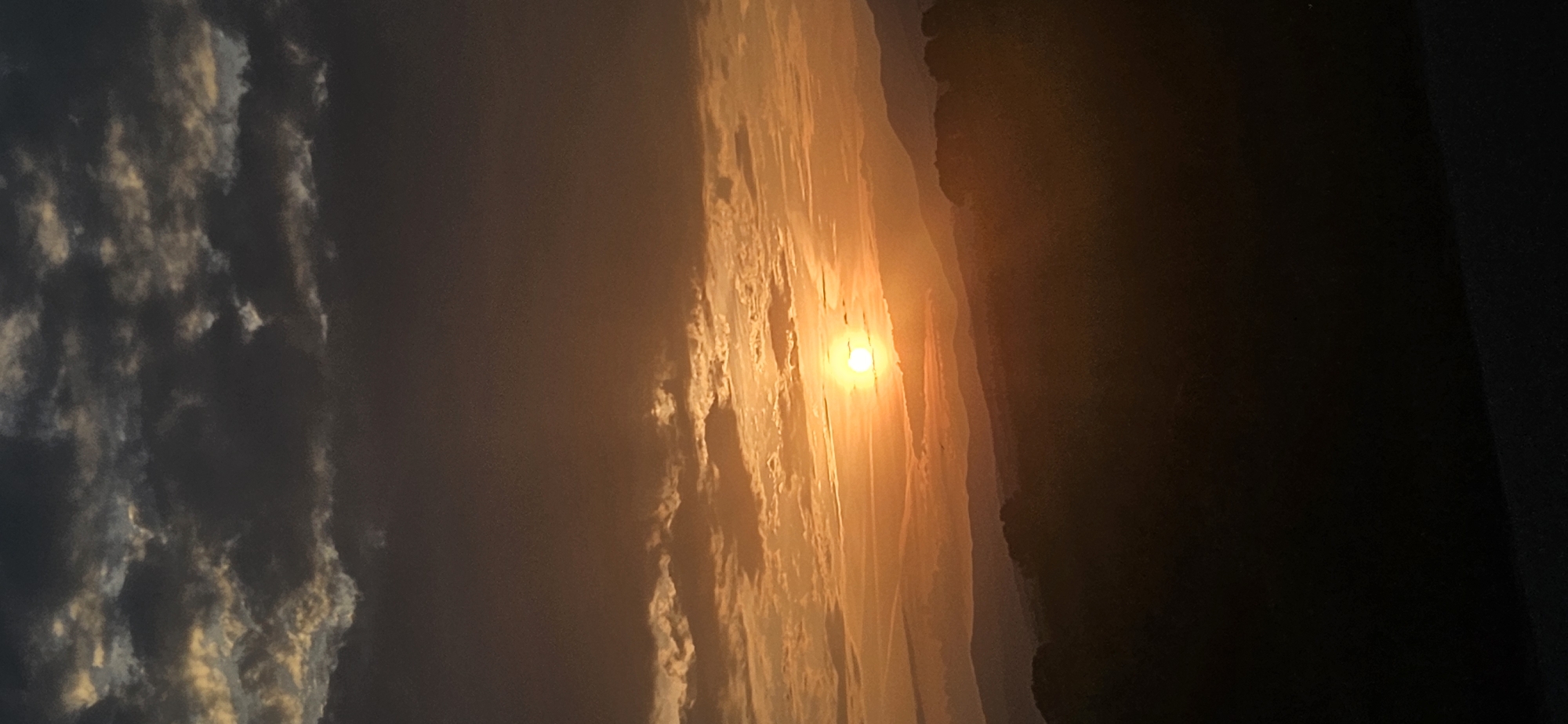 An outdoor, vertical format photograph captured at sunset in Momin Prohod, Bulgaria, depicts a panoramic view dominated by an intensely orange and yellow sun positioned slightly to the right of the central horizontal axis. The sun, a bright disc, is partially obscured by faint, horizontal cloud streaks, casting warm light across the mid-horizon. Above the sun, the sky transitions through shades of vibrant orange and yellow into a darker, muted brown and grey at the top, where dense, darker cumuliform clouds are visible, some with illuminated edges. Below the sun, a series of undulating mountain ranges or hills are silhouetted in progressively darker shades of blue-grey and purple, indicating atmospheric perspective. The immediate foreground is a dark, almost black silhouette of undulating terrain, likely dense vegetation or hills, creating a stark contrast with the illuminated sky and distant mountains. No people, discernible objects, or specific actions are visible in the scene. The overall impression is one of a tranquil, natural landscape during dusk.