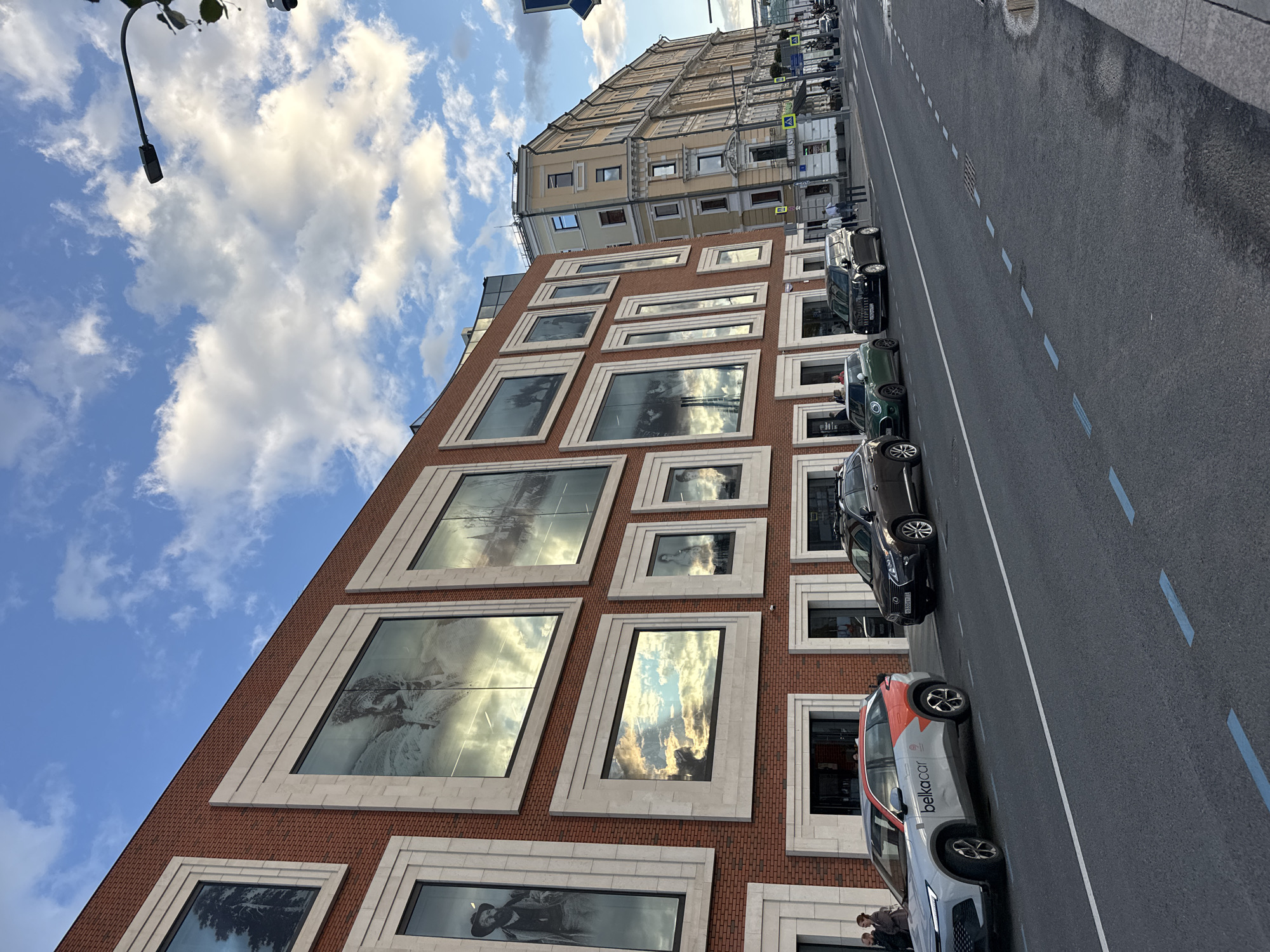 This is a street-level, eye-level, medium shot of a street in Moscow, Russia, featuring a modern brick building with large, framed windows on the left and older, light-yellow buildings stretching into the distance on the right. The sky is blue with scattered white clouds. In the foreground, there are several cars parked and driving on the asphalt street. The building on the left has a modern facade with large, rectangular windows that reflect the sky and clouds. Some windows display black and white images of people and trees. Below the windows are entrances with dark doors. The buildings on the right are older, multi-story structures with many windows, some with balconies. The street has white and blue lane markings. Pedestrians are visible on the sidewalks. A street lamp and a security camera are mounted on a pole on the right side of the image. The overall impression is an urban street scene during the daytime, possibly late afternoon or early evening given the light.