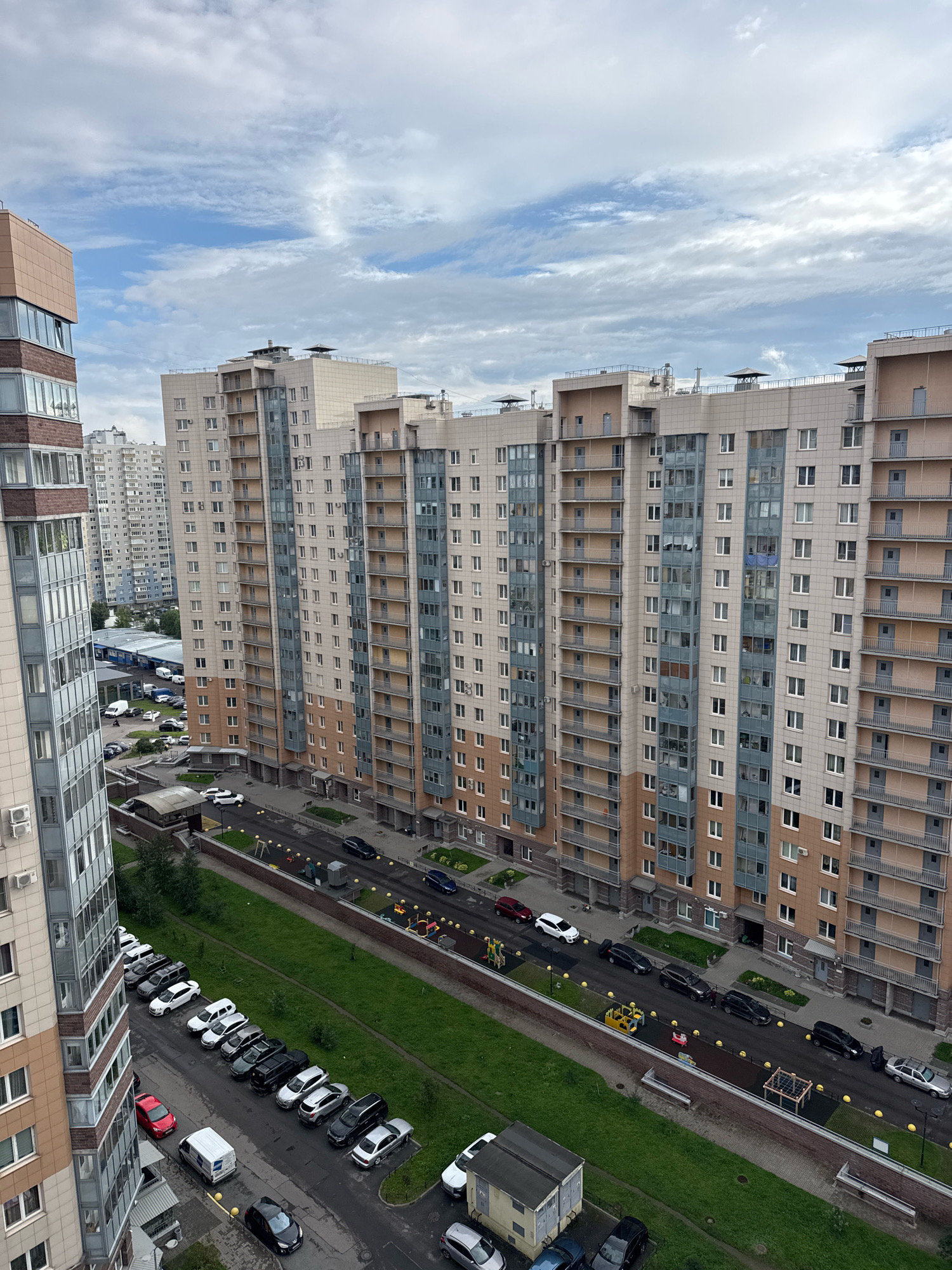 This is an eye-level, medium-distance, high-angle exterior shot of a residential area in an urban environment. The scene is dominated by a row of multi-story apartment buildings, characterized by their beige and gray facades, with numerous windows and balconies. Several cars are parked along the streets and in designated parking areas. Some of the vehicles are a mix of sedan and van types, with a variety of colors. A small playground area with yellow and red structures is visible. The sky above is overcast with scattered clouds. The overall environment appears to be a modern housing development with orderly landscaping and infrastructure.