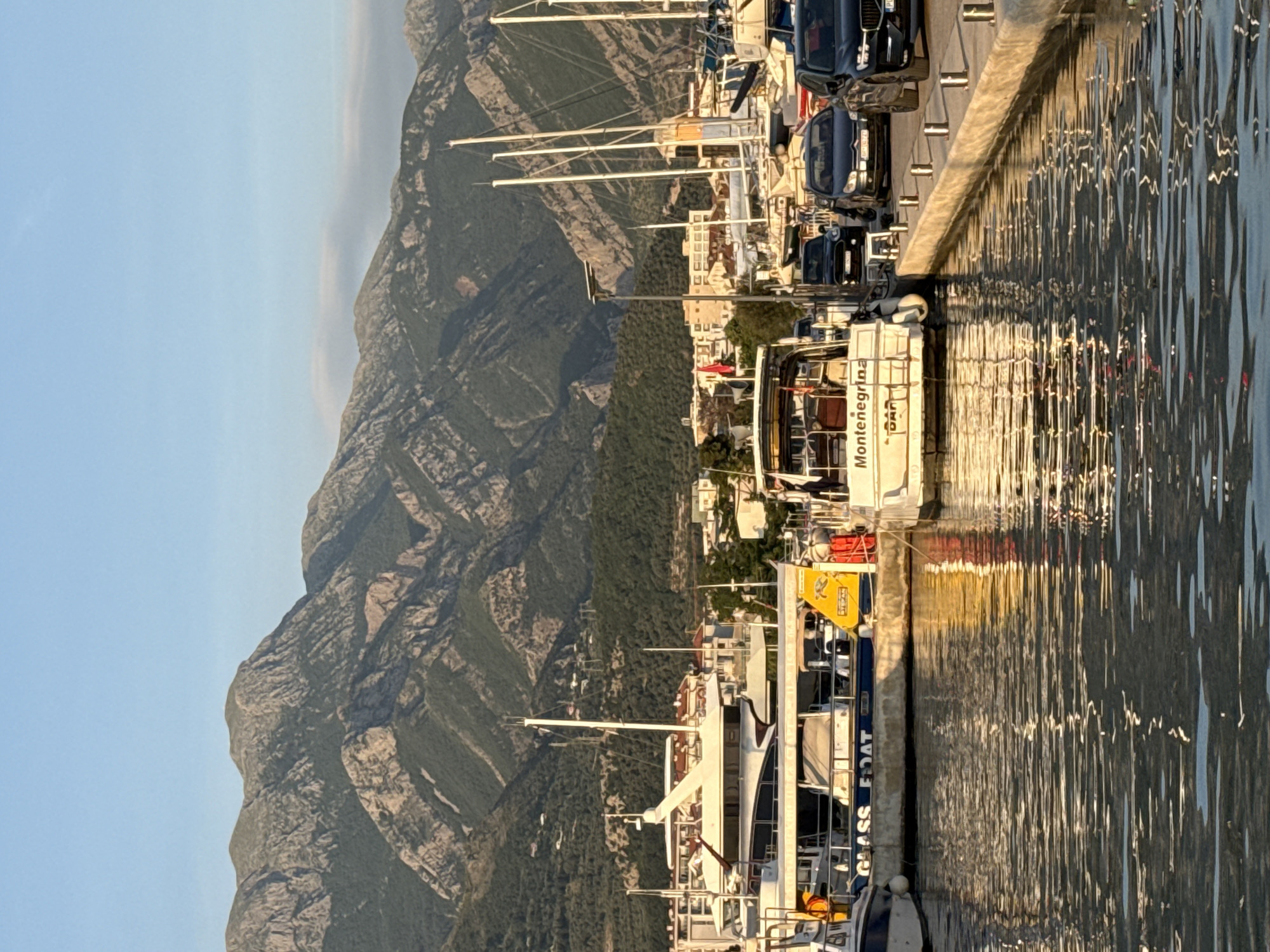 The image displays a picturesque harbor scene in Bar, Montenegro, captured during either late afternoon or early morning, as indicated by the warm, golden light reflecting on the water. The scene features a marina with numerous boats and yachts moored along a concrete quay.
In the midground, a white boat prominently displays the text "Montenegrina" and "BAR" on its hull. Closer to the foreground, another vessel has "GLASS BOAT" written on its side. Cars are parked along the quay behind the docked boats. The water in the foreground and midground is rippled, beautifully reflecting the sunlight and the structures of the boats and quay.
The background is dominated by a majestic and rugged mountain range, characterized by its rocky outcrops and patches of green vegetation. The mountains stretch across the horizon, providing a dramatic natural backdrop to the harbor. Above the mountains, the sky is clear and a soft blue, with a faint haze visible in the distance. The atmosphere is calm and serene, showcasing a harmonious blend of human activity in the marina and the imposing natural landscape. No specific individuals or activities are clearly visible.