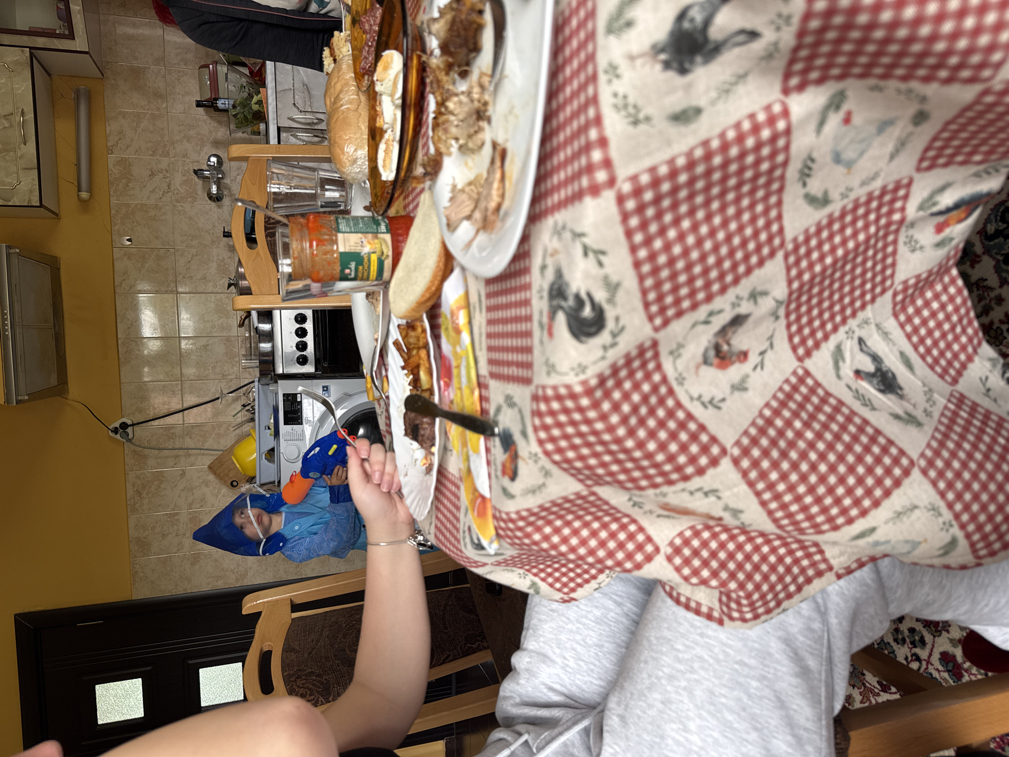 The image depicts an interior scene, likely a dining area or kitchen, with a table covered by a patterned tablecloth featuring red and white checks and images of chickens and birds. The table is laden with food, including platters of meat, bread, and condiments. A person is seated at the table, wearing gray sweatpants, with their arm extended towards the table holding a fork and a plate with yellow food. In the background, a child is visible wearing a blue costume with a pointed hat. The room includes kitchen appliances, cabinets, and tiled flooring. The location is Drenovets, Bulgaria.