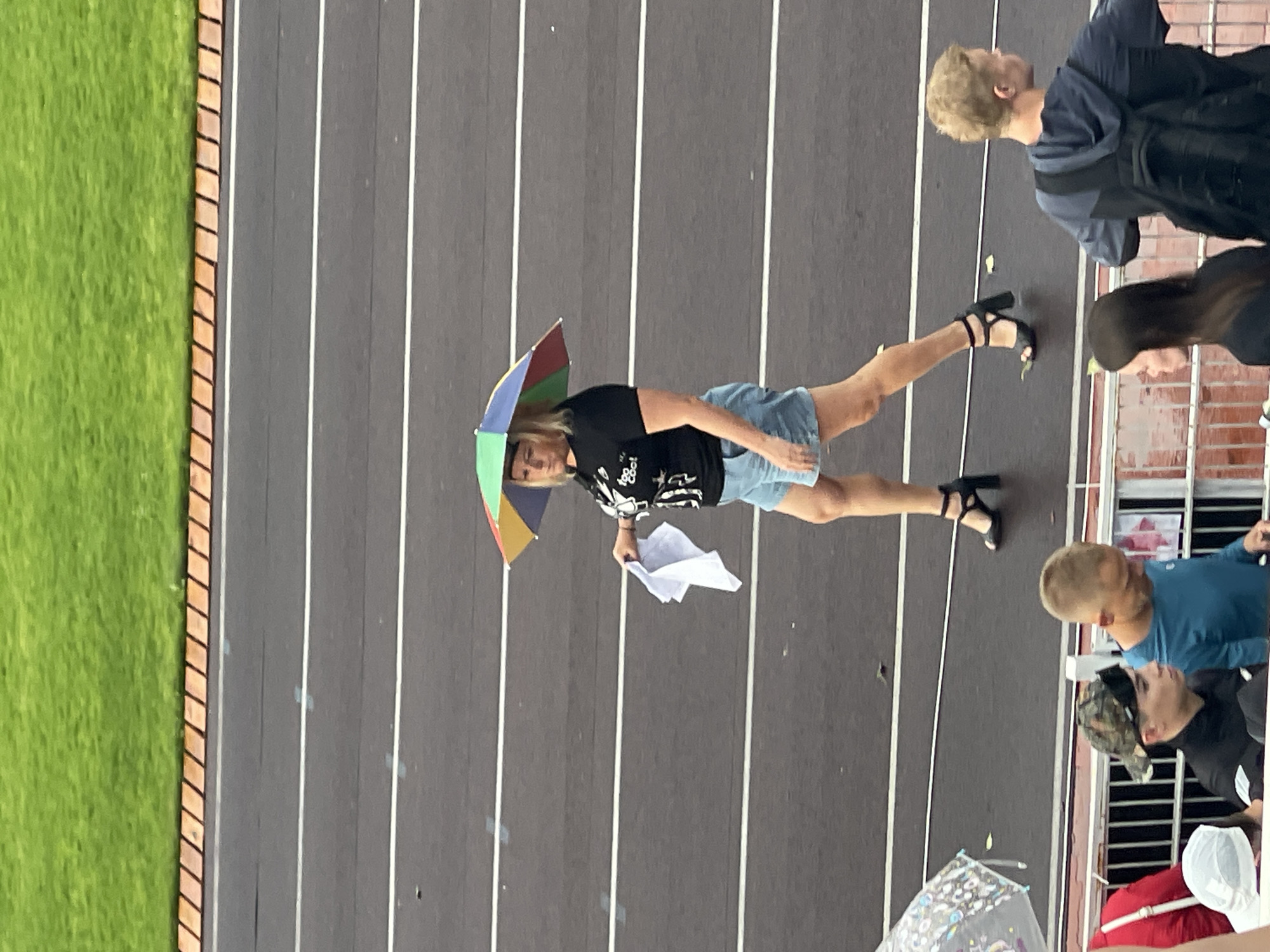 In an outdoor setting, possibly a sports track, a woman with blonde hair is walking. She is wearing a black t-shirt with a graphic design, light blue denim shorts, and black high-heeled shoes. A multi-colored umbrella is positioned above her head. In her right hand, she holds a folded white cloth. The background consists of a grey track with white lines and a grassy area beyond. In the foreground, other individuals are partially visible, suggesting a spectator area.