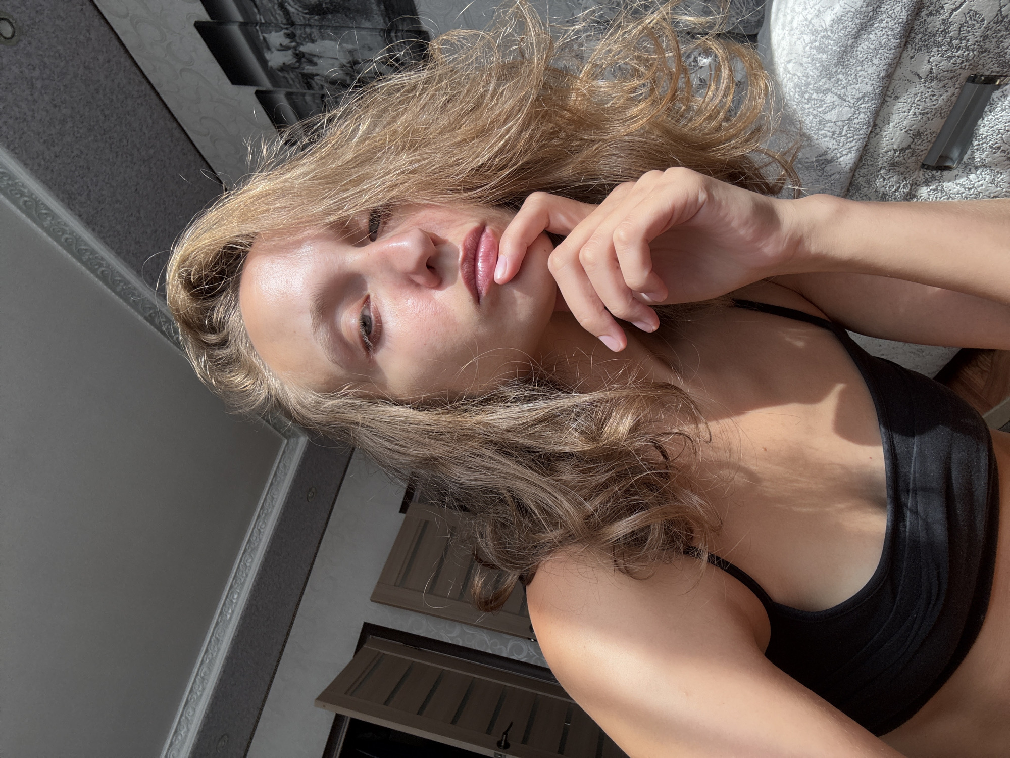 In a room with patterned wallpaper and a decorative ceiling border, a young woman with blonde, wavy hair is captured in a close-up, head-and-shoulders shot. Sunlight streams in from the left, casting dramatic shadows across her face and body, highlighting the texture of her skin and the waves of her hair. She is wearing a black top. Her right hand is raised, with her index finger resting thoughtfully on her chin. Her eyes are cast downwards, and her lips are slightly parted, suggesting a contemplative or relaxed pose. In the background, two doors are visible, one slightly ajar, and a framed artwork hangs on the wall. The scene appears to be indoors, possibly a residential space, with no clear indicators pointing to the specific city of Kuyeda, Russia.
