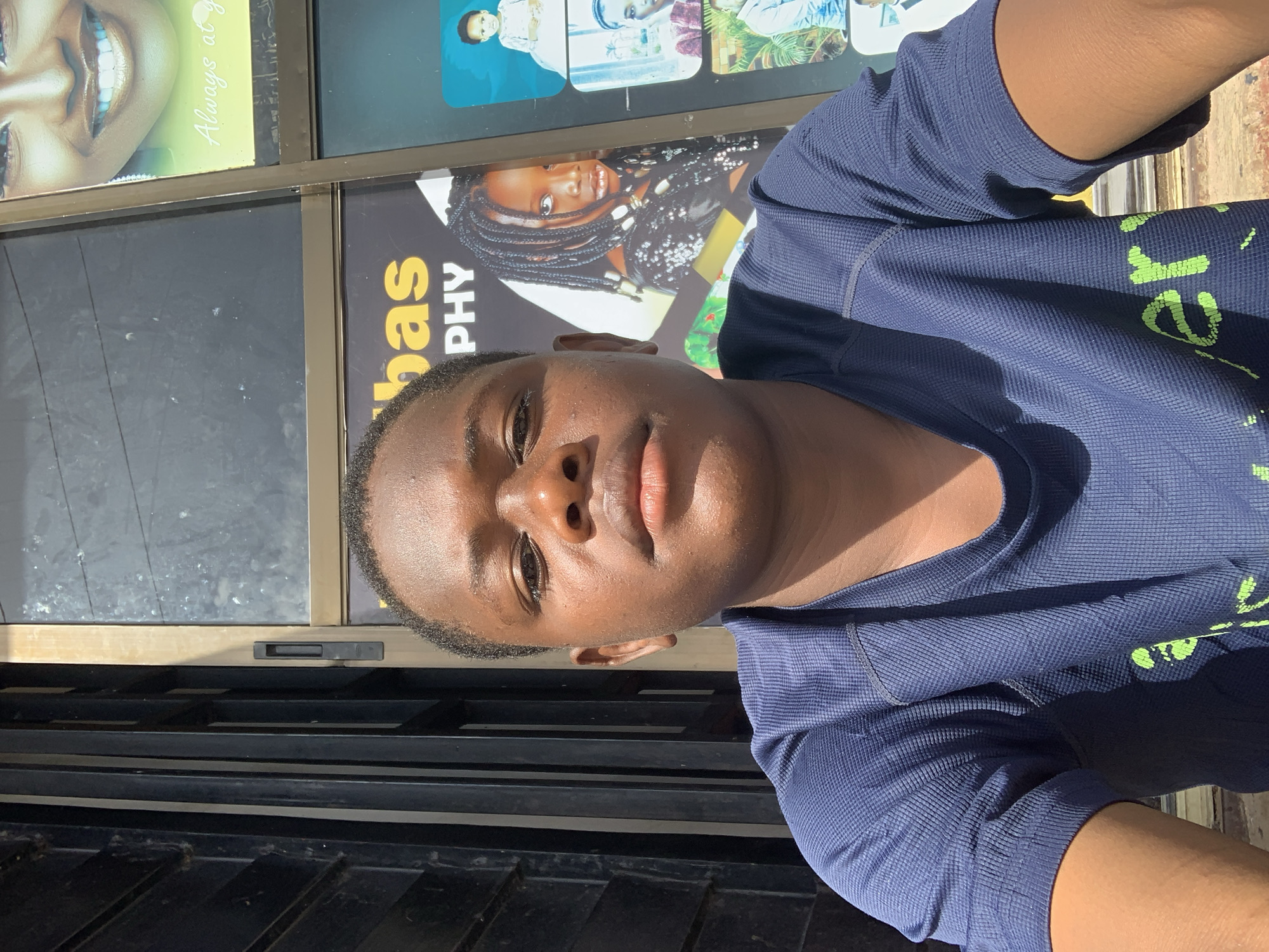 A young, dark-skinned individual is depicted in a close-up, chest-up portrait, directly facing the camera. The subject is wearing a navy blue, short-sleeved t-shirt with a textured, waffle-knit pattern. Partial bright green lettering, reading "ery," is visible on the lower front of the shirt. Direct sunlight illuminates the subject's face, particularly the forehead and nose, and the right arm, casting a shadow on the left side of the face and body.
The background on the left side of the image features dark, vertical and horizontal metal bars, indicative of a security gate or barrier. The background on the right and directly behind the subject consists of a glass window or door framed in a light-colored material. Through this glass, multiple commercial posters are visible. A prominent poster behind the subject's head displays large yellow text reading "bas" and, beneath it, "PHY," strongly suggesting a photography business. Adjacent to this text, a portrait of a young girl with braided hair is visible, looking towards the viewer. To the upper right, a partial poster shows the lower face of a smiling woman with white teeth, accompanied by yellow script text that reads "Always at y." Below this, a grid of smaller framed images includes portraits of a baby in a white outfit, a young man in a patterned shirt, and another man in a light shirt, all appearing to be studio photographs.
The scene portrays an outdoor setting in front of a commercial building, specifically a photography studio, located in the city of Jos, Nigeria.