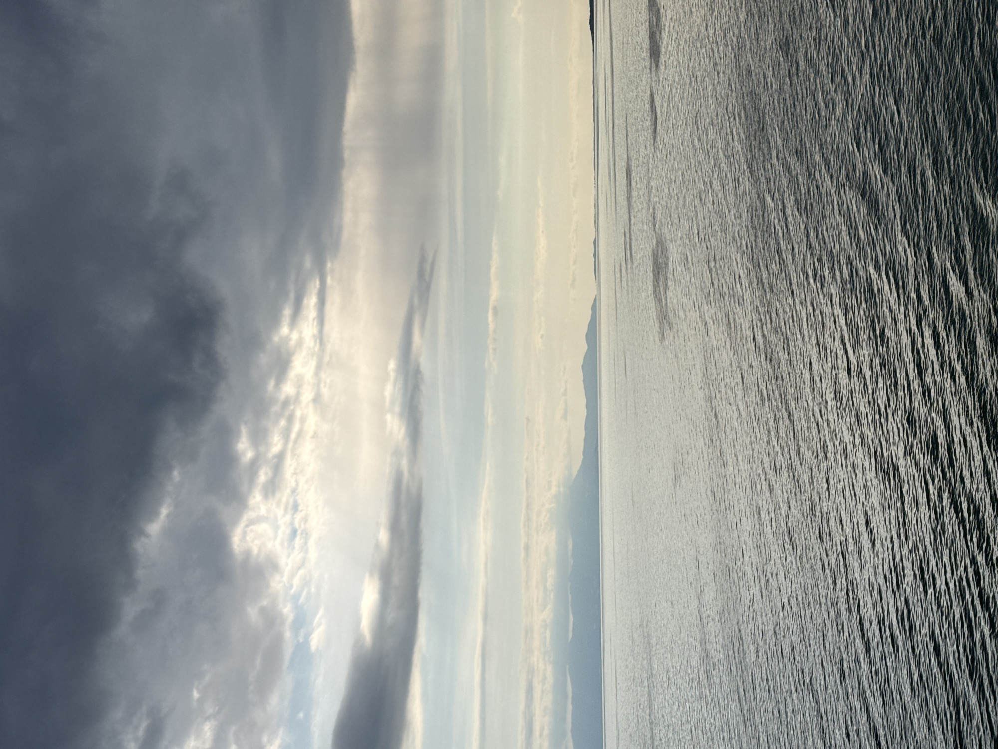This landscape photograph, taken outdoors in the city of Fanari, Greece, presents a study in contrasts between the sky and the sea. The left portion of the frame is dominated by a dynamic sky filled with dark, voluminous clouds. These clouds are illuminated by diffused sunlight. A stretch of sky follows, that consists of various streaks of white and light gray clouds. A thin line of land, likely a distant coastline, is visible across the middle. The right portion of the image is occupied by the sea, its surface marked by intricate patterns of waves, creating a textured, light gray and dark gray appearance. The light reflects off the water's surface, emphasizing the movement and the grainy texture of the water.