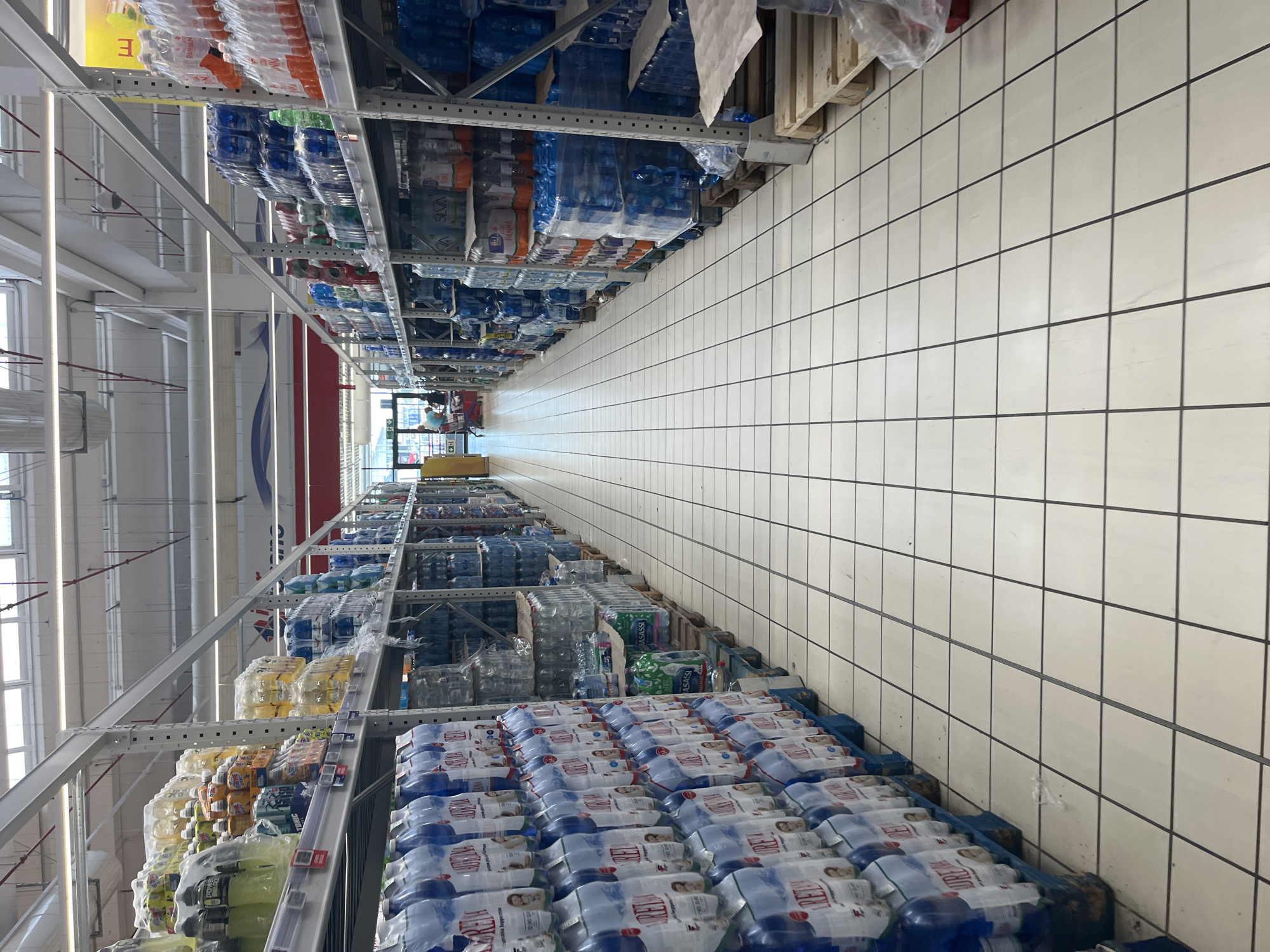 The image shows the interior of a supermarket aisle in Castiglione delle Stiviere, Italy, with shelves stocked with bottled water and other beverages. The flooring is tiled in a light color with dark grout lines. The aisle is wide and appears well-lit, with overhead fluorescent lighting. On the left side of the aisle, tall shelves are filled with cases of bottled water, many of which are shrink-wrapped. Some of the bottles have labels visible, with prominent brands and colors. On the right side, similar shelving units are also stocked with cases of bottled water, as well as some other types of drinks in bottles and multipacks. Pallets are visible at the base of some shelves, holding additional stock. In the distance, at the end of the aisle, two individuals are partially visible near a shopping cart, one of whom appears to be a shopper and the other a supermarket employee. The environment is clean and organized, characteristic of a retail grocery store.