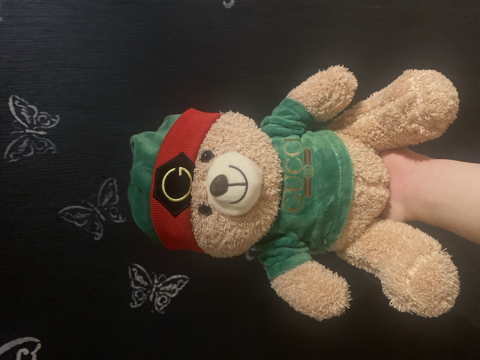 A light brown teddy bear, dressed in a green shirt with the "GUCCI" logo and a red headband with a black and gold emblem, is held up by a hand in front of a dark background. The background is a black wall with white chalk drawings of butterflies in various sizes.