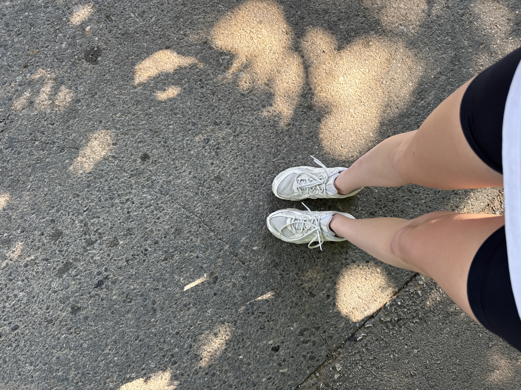A top-down shot shows a person standing on a grey asphalt surface, presumably a sidewalk or street. The person is wearing white sneakers, black athletic shorts, and a white shirt (only partially visible). Sunlight filtering through trees creates dappled shadows on the ground. The location is an outdoor area, likely in a city.