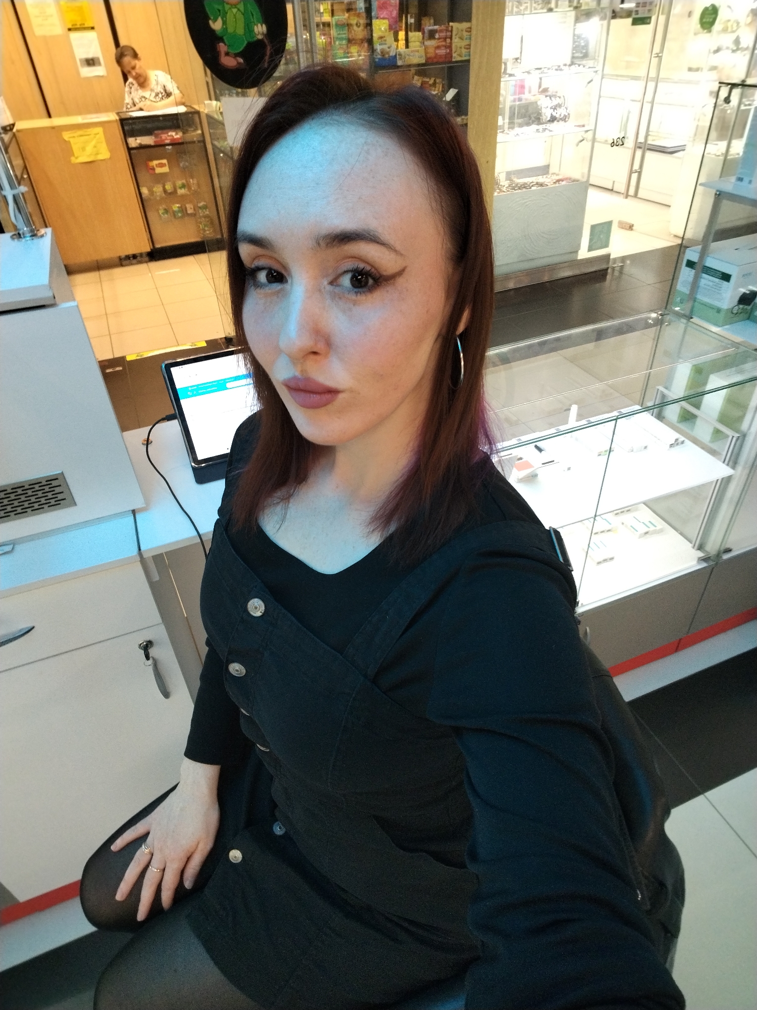 A young woman with reddish-brown hair, distinct winged eyeliner, and light lipstick is the central subject of this indoor photograph, looking directly at the camera with a poised expression. She wears a black long-sleeved top underneath a dark, possibly denim, pinafore dress with light-colored buttons, and hoop earrings. Her right hand, adorned with two rings on her ring finger, rests on her knee, visible above black tights.
The setting appears to be a commercial establishment in Izhevsk, Russia, possibly a shop, kiosk, or a corner of a larger store. To the main subject's left, a white counter holds a tablet displaying a light blue interface, connected by a black cable. Below this counter, keys hang from a lock on a white cabinet.
In the background, an older woman with light hair and a patterned top is seated at a wooden counter, seemingly occupied with something on its surface. Above her counter, shelves display various products, and a green circular sign featuring a cartoon character is visible. To the right of the main subject, several glass display cases exhibit small items, suggesting merchandise like jewelry or accessories. The floor is composed of light-colored tiles. The scene is brightly lit by artificial indoor lighting. No distinct, legible text is visible on any signs, though reflections on a glass case are present.