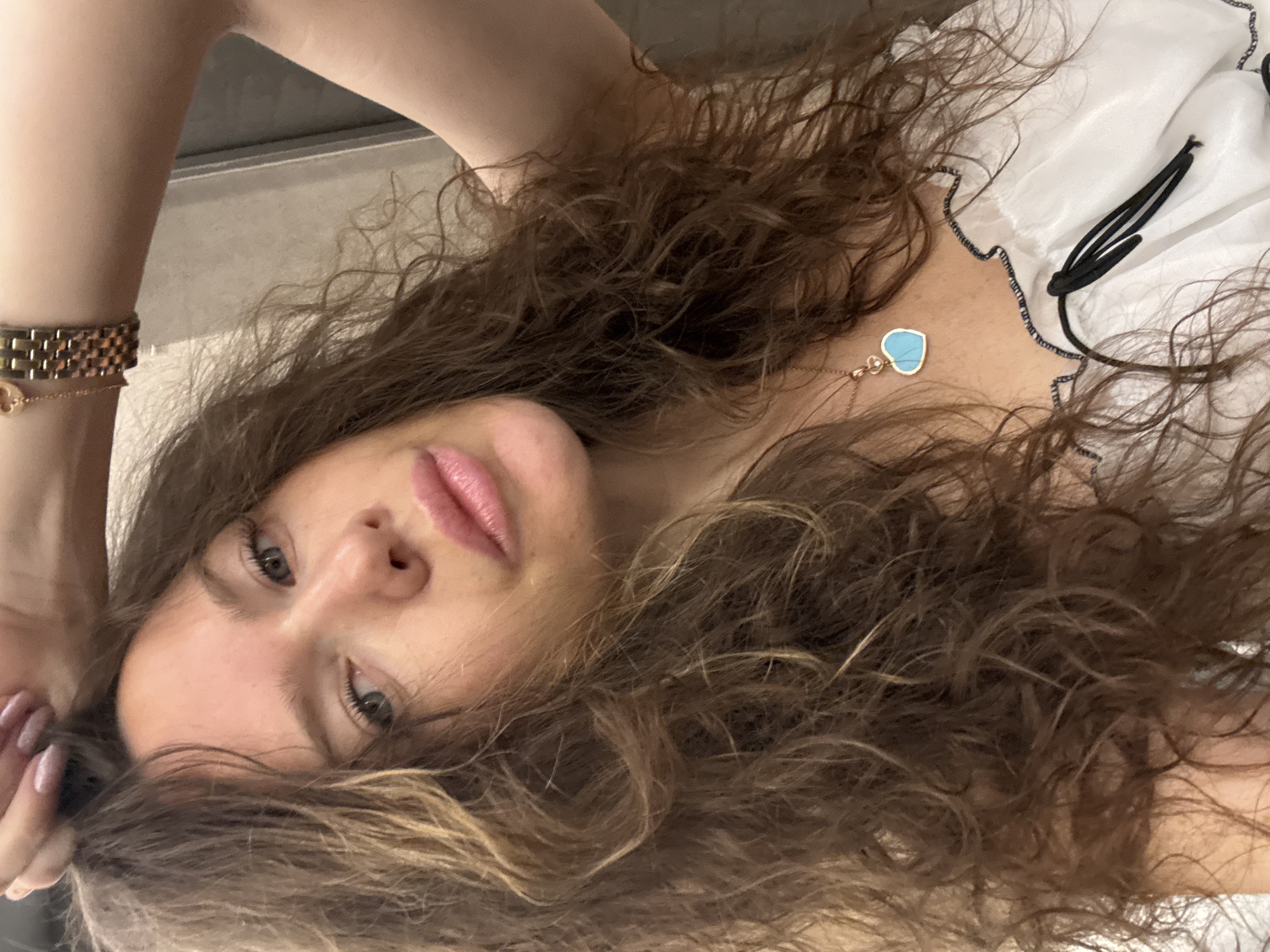A close-up portrait shows a young woman with long, wavy brown hair, lying down and looking directly at the camera. Her head is angled, with her face occupying the lower-left section of the frame. She has light eyes and her lips are slightly parted.
She is wearing a white top with black decorative trim along the neckline and a possible black tie detail. A delicate gold-toned necklace with a light blue, heart-shaped pendant and a small clear stone accent rests on her chest. On her raised left wrist, she wears a gold-toned bracelet with rectangular links and a finer gold-toned chain bracelet with a small heart charm. Her fingernails, visible on her left hand near her face, have light pink polish.
The background consists of a soft, light-colored surface, likely indicating an indoor setting, with an indistinct darker area in the upper left. No specific location cues for Khimki, Russia, or any text are visible in the image. Her expression is neutral and direct.