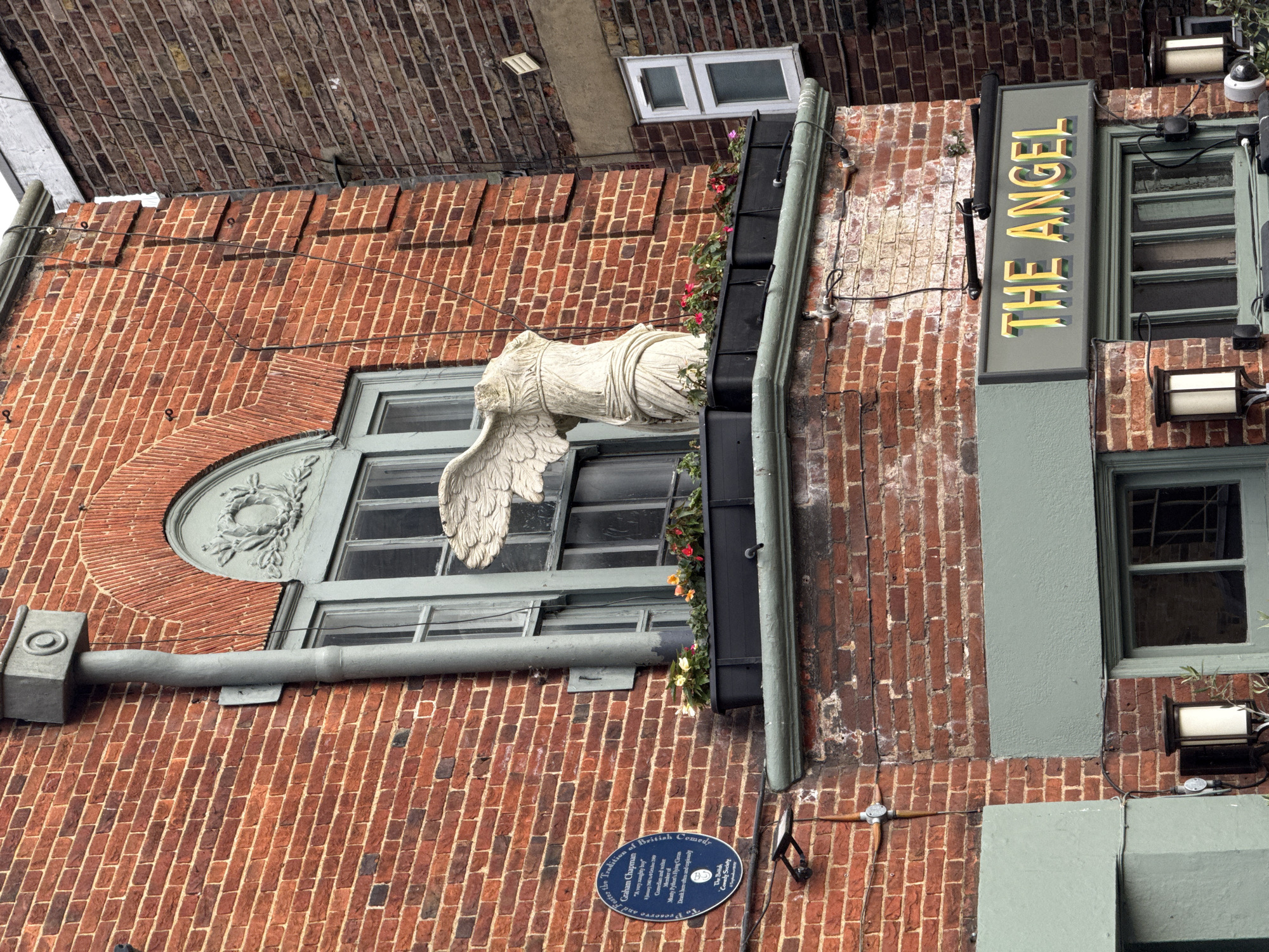The image shows the exterior of a red brick building, presumably a pub called "The Angel," as indicated by the vertical sign with the name in bold yellow letters. A partial angel statue, consisting of a torso and wing, is mounted above a window on the brick facade. The window itself has a decorative molding above it.
To the lower right of the window is an oval plaque dedicating the space to Graham Chapman, a member of the British comedy group, "Monty Python's Flying Circus." The plaque also indicates the establishment is dedicated to preserving the traditions of British Comedy. Additional details include a black gutter system with a flower box, lighting fixtures mounted on the brick, and a view of the sky obscured by the building's upper structure. The building is in London, United Kingdom.