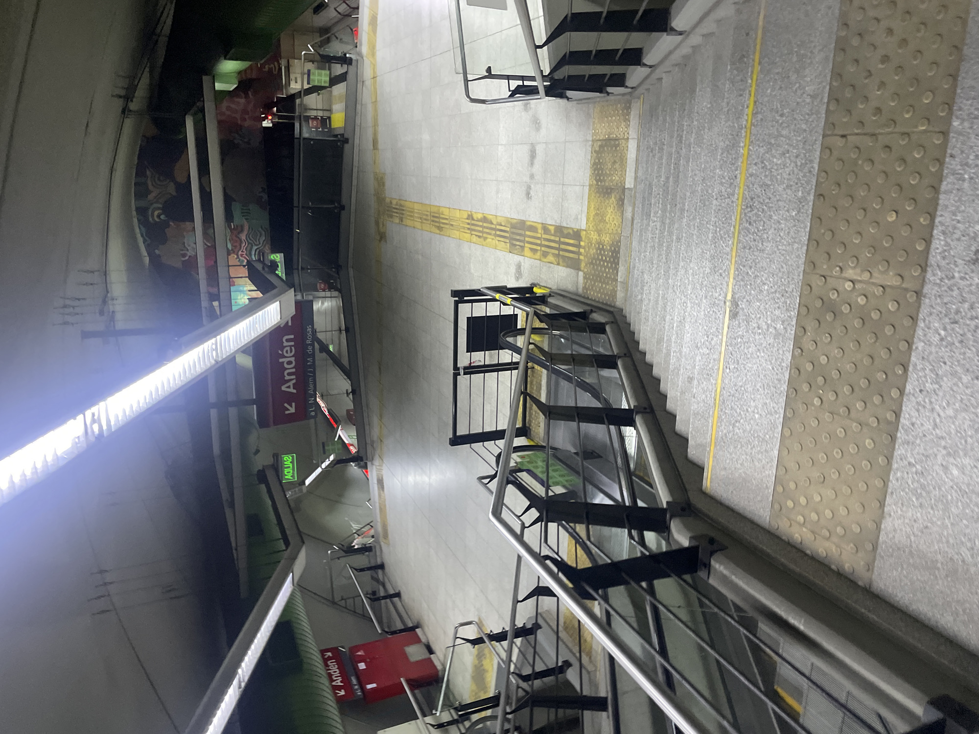 The image shows an interior view of a subway station in Buenos Aires, Argentina. There are staircases with metal railings leading downwards. The floor has tactile paving and a yellow guiding line for the visually impaired. Signage is visible indicating the direction to "Andén" (platform). There's a mural or artwork visible on the wall of the subway tunnel. The general environment is typical of a subway station with artificial lighting.