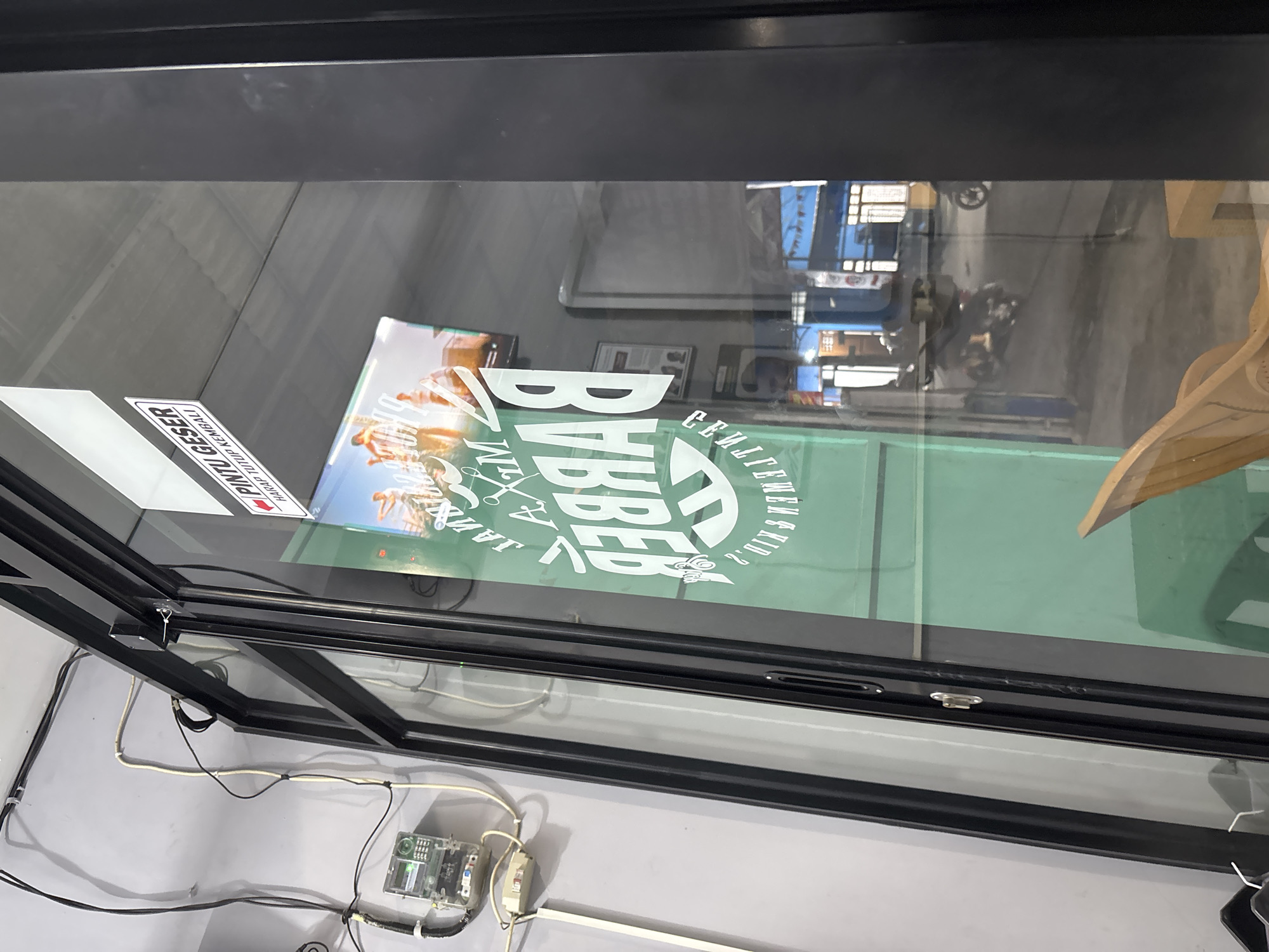 This is a close-up, low-angle, eye-level shot of the entrance to a barber shop in Wanajaya, Indonesia. The shop's glass doors are slightly ajar, revealing a glimpse of the interior and the street outside. A sign on the door reads "PINTU GESER" (Sliding Door) and "HARAP TUTUP KEMBALI" (Please Close Again). Inside, a large, stylized logo for "MR. BARBER" is visible on the green glass. The street scene shows motorbikes and shopfronts. Electrical conduits and an electricity meter are mounted on the grey wall to the left of the entrance. A wooden chair is visible in the lower right corner.