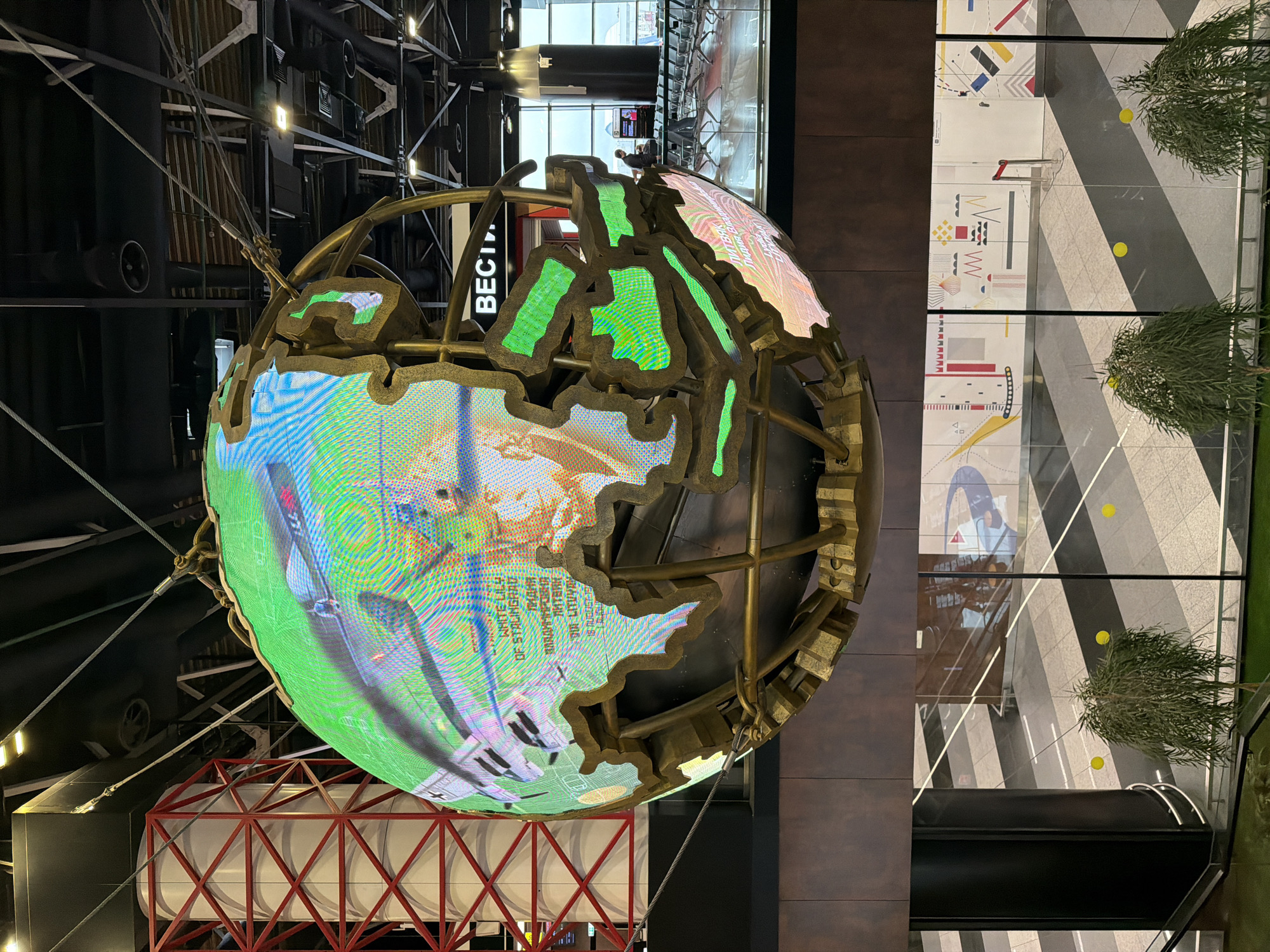 Here is a description of the image:
The image shows an indoor installation featuring a large globe with a digital display. The globe is encased in a bronze-colored framework resembling the outline of continents, with green lighting highlighting these outlines. The digital display shows a variety of images, including what appears to be aviation-related content like airplanes and text, with bright green and blue hues dominating.
The globe is suspended from the ceiling by cables and is situated above a red and white structural support. The ceiling itself is a complex network of pipes, beams, and lighting fixtures.
To the right of the globe is a wall of large glass panels. The panels reflect images from the interior, including geometric patterns on the floor, plants, and lighting.
There are no people visible in the immediate foreground of the image. The scene appears to be inside a museum or exhibition hall.