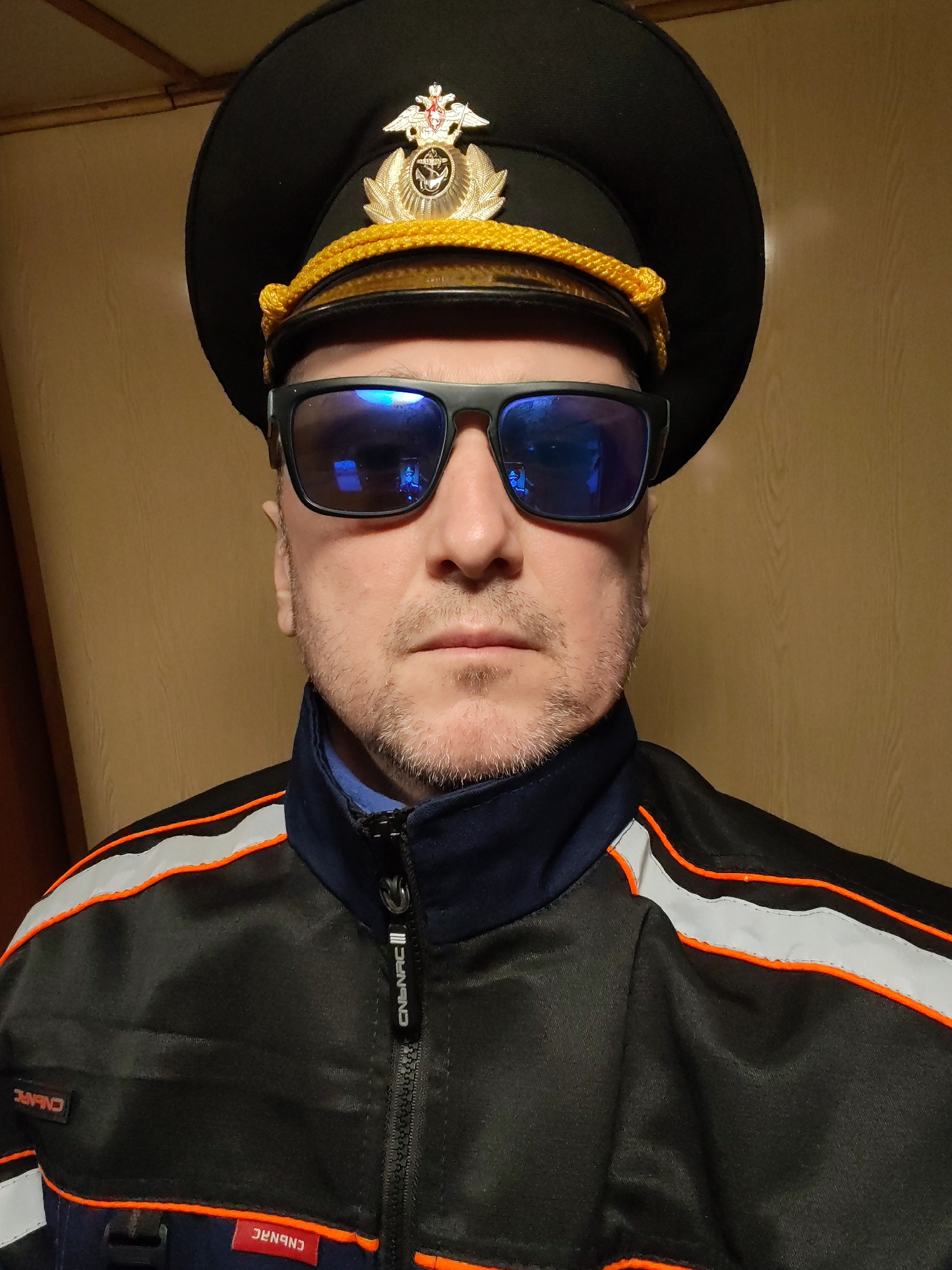 A man is depicted in a close-up, chest-up portrait indoors. He has light stubble or a short beard and is wearing dark sunglasses with blue reflective lenses, obscuring his eyes. He is wearing a black uniform-style cap with a golden braided cord across the front and a prominent golden insignia at the center. The insignia features a double-headed eagle holding a shield, suggestive of a Russian state emblem.
The man is also wearing a black jacket with a navy blue collar and inner lining. The jacket has distinct orange piping and a wider grey reflective stripe running horizontally across the chest and shoulders. The jacket features a zipper closure down the front.
Visible text on the jacket includes "CNBNRC" on a small red tag on the lower left, and the same text "CNBNRC" (possibly "CNBNRC |||") vertically on the zipper pull, which appears to be a brand name. Another mirrored "CNBNRC" is visible on a tag near the left shoulder.
The background is a plain, light brown or tan wall, possibly with a subtle wood grain texture, providing a simple and uncluttered setting for the portrait. The lighting suggests an indoor environment. The man has a neutral expression, looking directly at the viewer. There is no specific activity taking place beyond posing for the photograph.