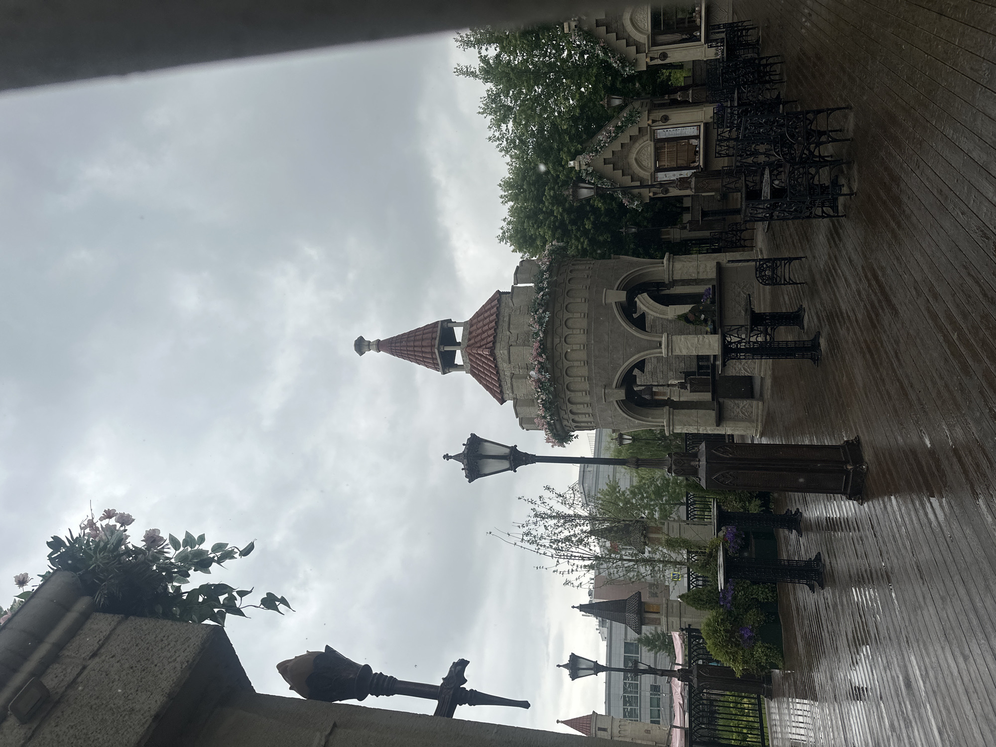 The image depicts an outdoor seating area with a medieval theme. The ground is made of dark wooden planks, which are wet, suggesting recent rain. Several black metal tables and chairs are arranged on the deck. In the background, there are structures resembling small castles or towers with red tiled roofs. One prominent circular tower has decorative floral arrangements around its top. There are also smaller, gabled buildings that appear to be kiosks or food stalls. Traditional lampposts are visible, adding to the atmosphere. The sky is overcast, indicating a cloudy or possibly rainy day. The scene appears to be a recreational or dining area, possibly part of a park or entertainment venue. The location is the Gorky Park area in Moscow, Russia.