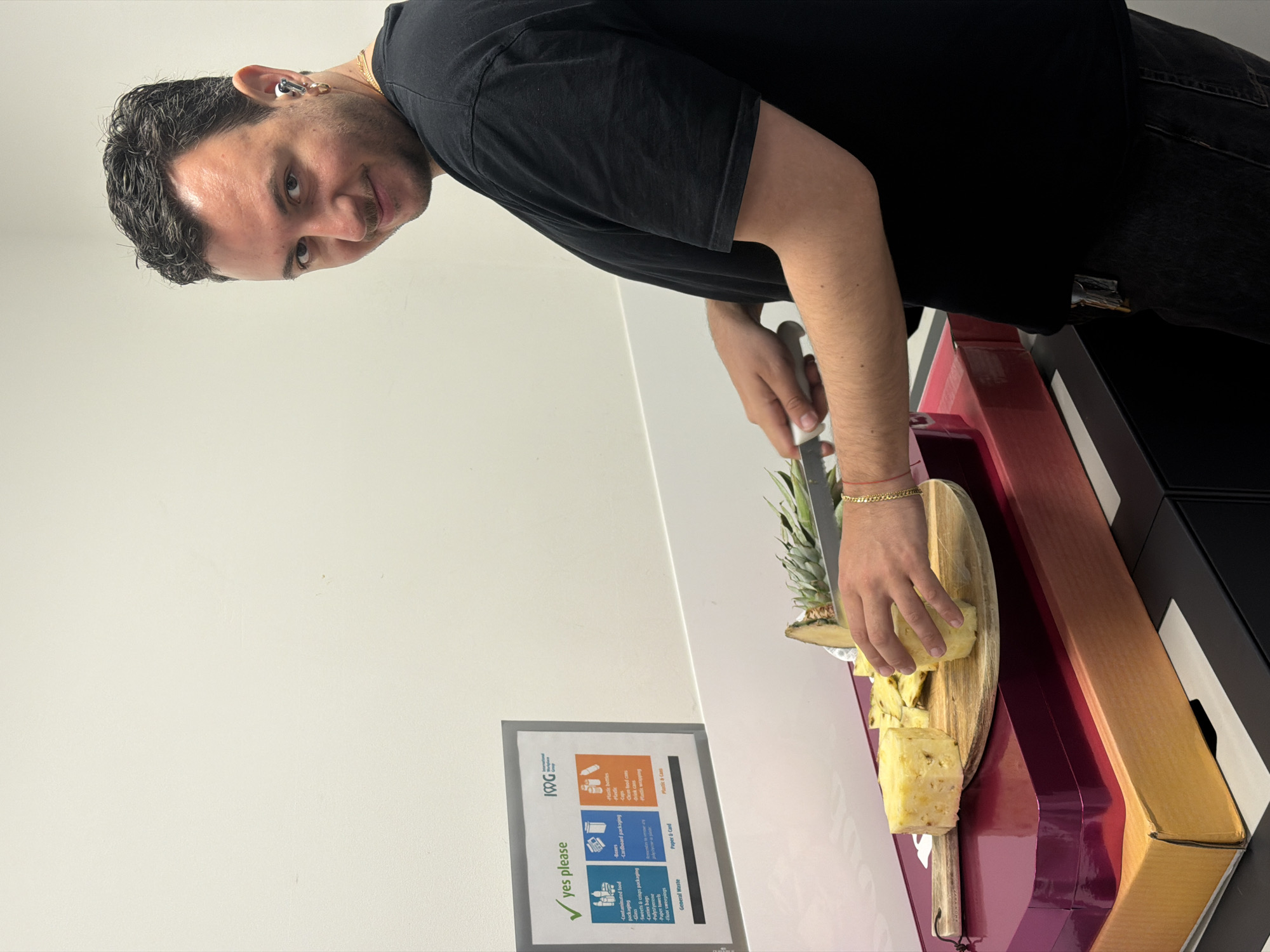 A slightly angled, medium shot depicts a man cutting pineapple on a wooden cutting board. He is positioned in front of a light-colored wall featuring a recycling information sign labeled "yes please" with graphics illustrating different waste categories, under the logo of "IOGG." The man, facing the camera, has dark hair, a light complexion, a gold earring, and a gold bracelet. He is wearing a black t-shirt. He uses a knife held in his right hand to cut the pineapple, which is sitting on the wooden cutting board. A red and gold box is visible next to the cutting board. The location appears to be indoors, in the City of London, United Kingdom.