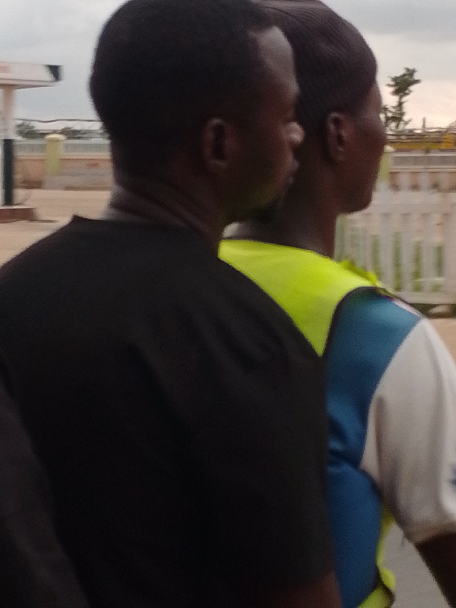 The image shows two individuals in Gabukka, Nigeria. The person in the foreground on the left is wearing a black t-shirt. To their right, and slightly behind, is another person wearing a bright yellow and blue vest over a white shirt. The background reveals a street scene with what appears to be a gas station and a white fence. The sky is overcast. The overall scene suggests an outdoor setting in a moderately urban or semi-urban area.