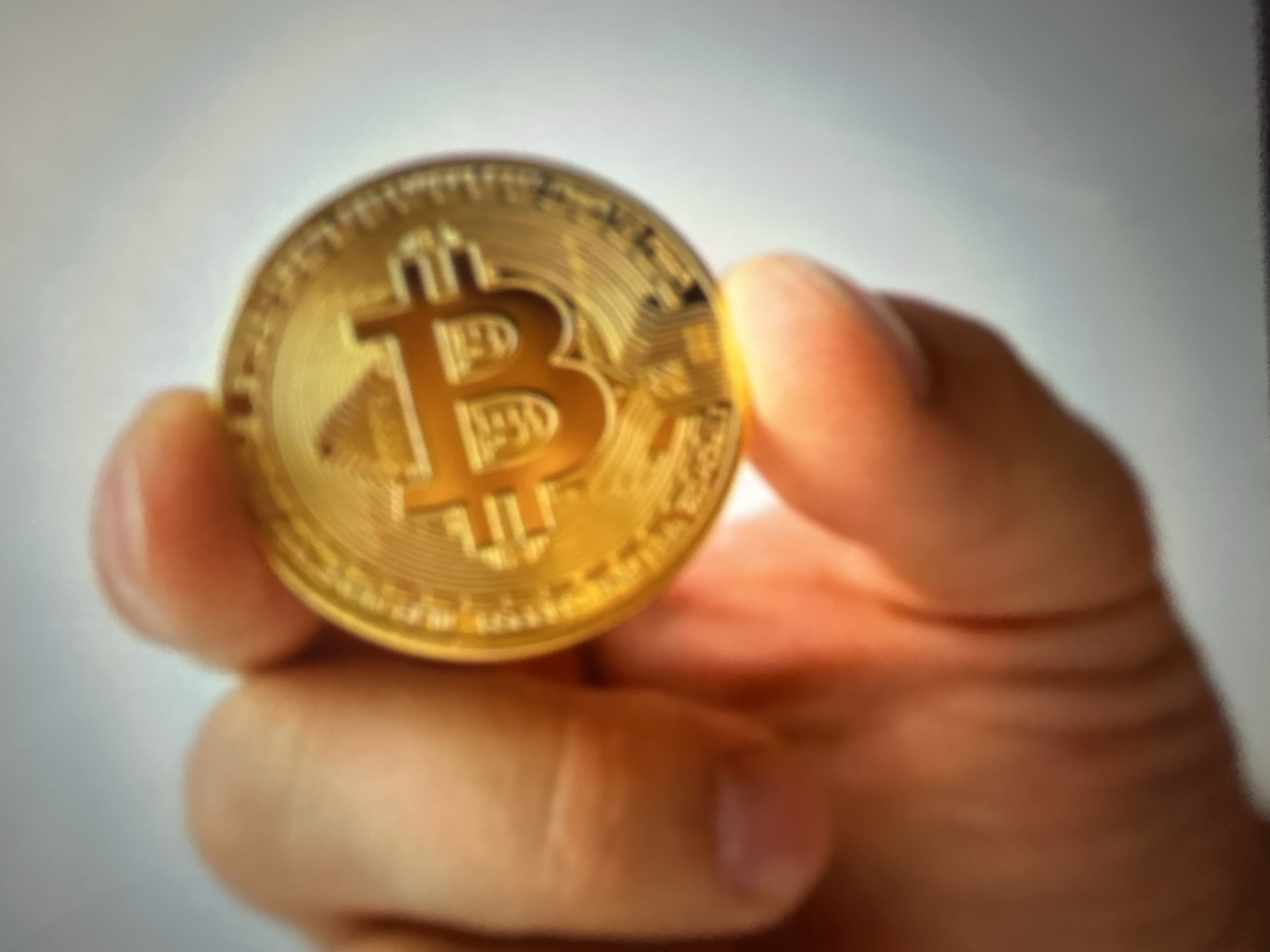 The image shows a close-up of a hand holding a gold-colored coin with a large "B" embossed on it, which is a symbol for Bitcoin. The coin is golden and appears to be a replica of a Bitcoin cryptocurrency. The hand is holding the coin between the thumb and index finger. The background is blurred and appears to be a light-colored, out-of-focus surface. The lighting suggests an indoor setting, with a bright light source illuminating the coin and the hand, creating some glare. The image is slightly out of focus, which gives it a soft, abstract quality. There is no visible text on the coin in this particular image. The context of Gelendzhik, Russia, is not visually evident from the image itself.