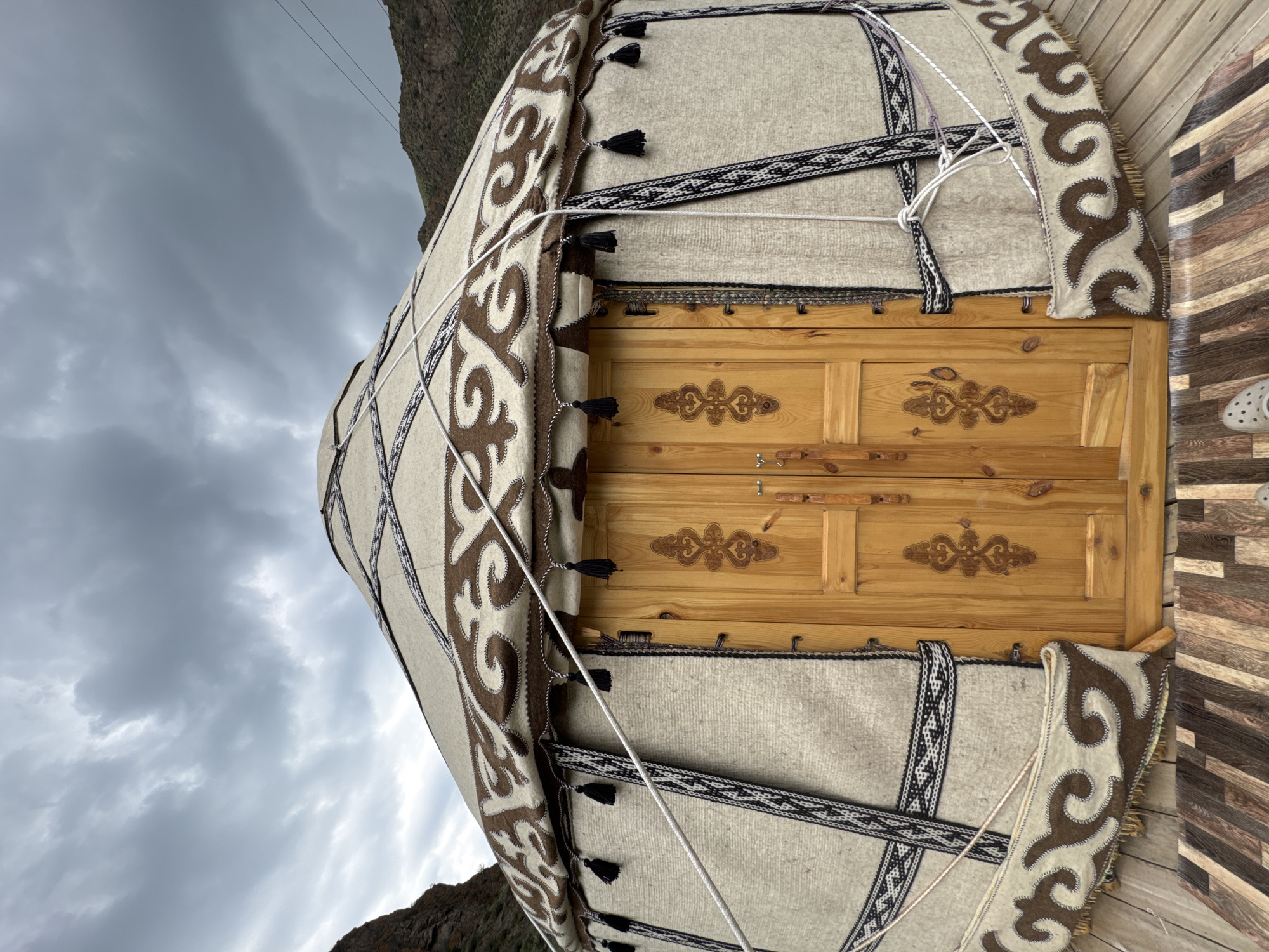 The image shows a traditional yurt, a portable dwelling, set against a backdrop of cloudy skies and mountainous terrain. The yurt is made of felt and fabric, adorned with intricate patterns in brown and white. A wooden door with decorative carvings is centrally located on the yurt's exterior. Ropes are used to secure the yurt, and tassels hang from the upper portion of the structure. The yurt is positioned on a wooden platform, suggesting it is part of a campsite or tourist facility. The scene appears to be in Kyzyl-Korgon, Kyrgyzstan, indicated by the context. No people are visible in the frame. The environment is natural and rugged, consistent with a rural or mountainous region.