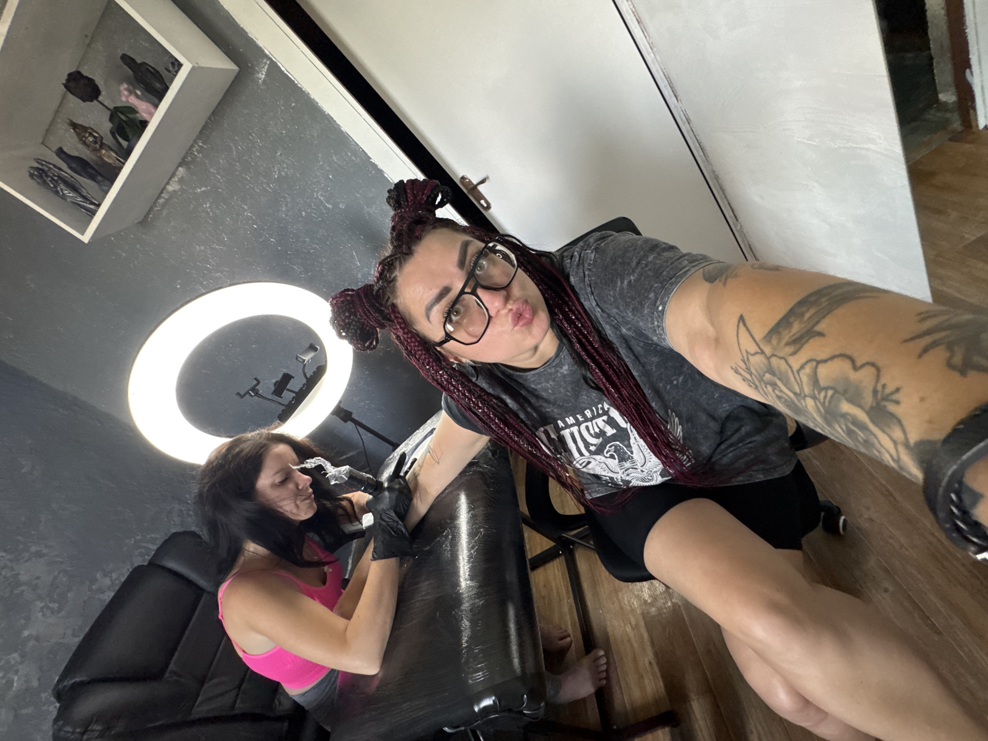 The media file shows two individuals in what appears to be a tattoo studio. One person, with dark hair styled in braids and wearing glasses, is taking a selfie. They are wearing a gray t-shirt with "AMERICA" and an eagle design printed on it, and black shorts. Their arm is heavily tattooed. The second person, with long dark hair and wearing a pink tank top, is focused on applying a tattoo to the first person's arm, wearing black gloves. A bright ring light is positioned behind them, illuminating the scene. The room has a dark gray textured wall and wooden flooring. A shelf with several decorative items, possibly ceramic figures, is visible in the upper left corner. The overall atmosphere suggests a professional tattooing session in progress, possibly during daylight hours given the bright lighting. The exact time of day and weather are not discernible.
