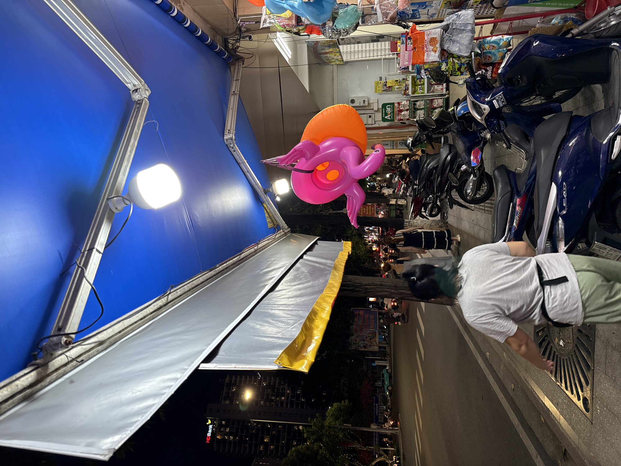 This is a nighttime street scene in Nha Trang, Vietnam, likely on a sidewalk in front of a shop. A blue awning covers the storefront, illuminated by a bright overhead light and possibly lights from within the shop. A woman with dark hair, wearing a gray t-shirt and light green pants, walks away from the camera on the sidewalk. She has a black sash tied around her waist. In front of her, a large metal grate is visible in the sidewalk. Beyond the woman, a street with traffic, including motorcycles and cars, is visible. The background shows buildings, trees, and streetlights. Hanging from the awning is a large, inflatable pink octopus toy with an orange ring. The shop to the right displays various items on shelves and racks, including bags of snacks, clothing items, and plastic containers. Several motorcycles are parked along the sidewalk. The overall atmosphere suggests a busy urban environment at night.