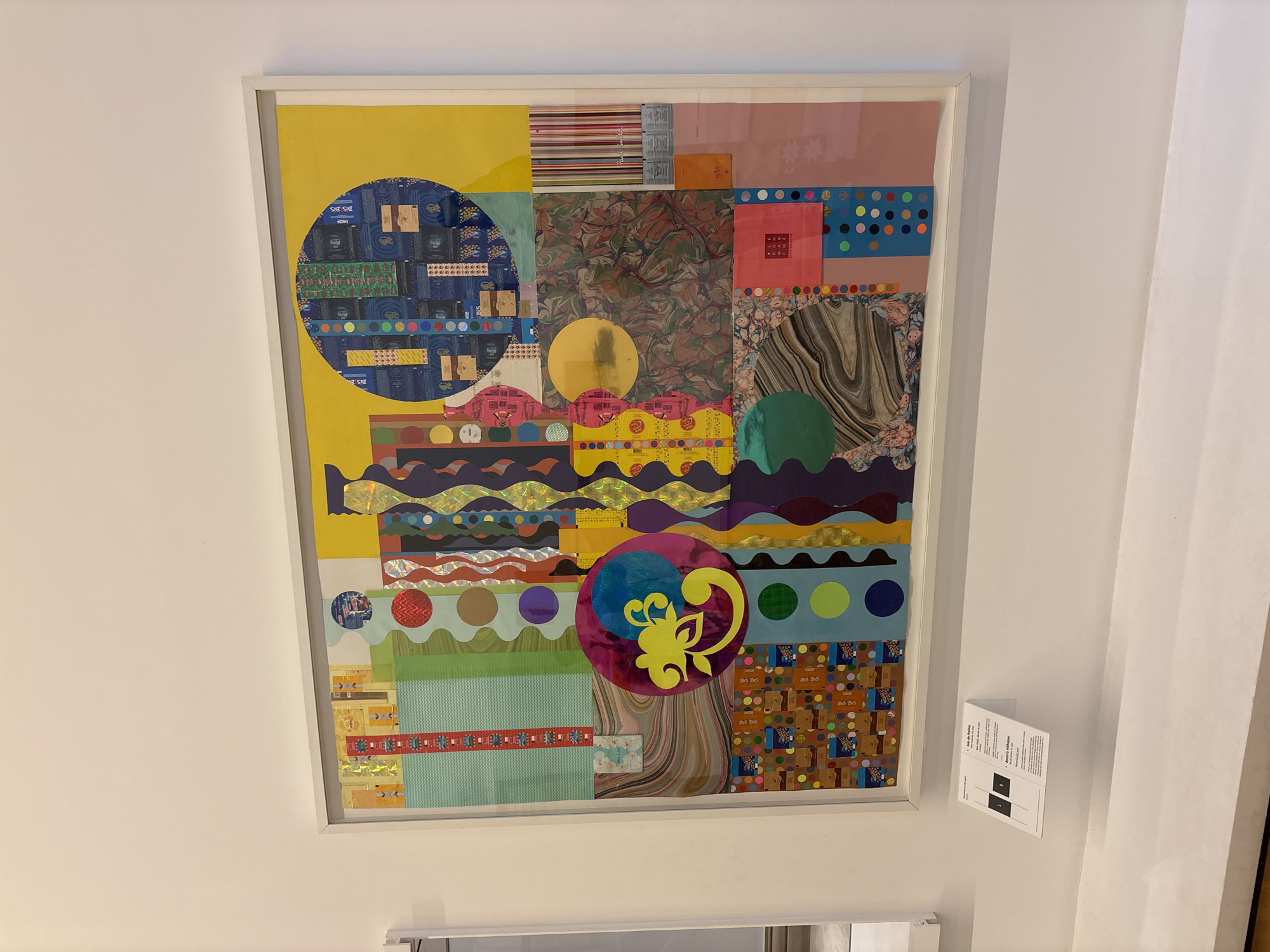 A framed abstract collage hangs on a white wall. The collage incorporates various materials and patterns including colorful circles, stripes, floral patterns, wavy lines, and geometric shapes. The colors range from yellow and pink to blue and green. A small white card is pinned to the wall beside the framed collage. The scene is located in the city of São Paulo, Brazil.