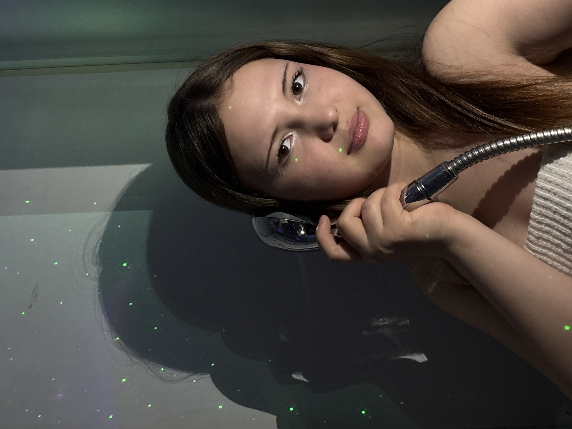 A close-up portrait of a young woman is presented with a 90-degree clockwise rotation, depicting her seemingly lying down on her back, holding a shower head. She has long, dark brown hair that cascades around her. Her face is turned towards the viewer, featuring light skin, brown eyes, and plump lips with visible lip gloss. She wears subtle makeup, including white eyeliner on her lower lash line.
She holds a metallic, flexible shower hose and head with her right hand, resting it near her ear. A light-colored, possibly striped, fabric, resembling a towel or clothing, is visible over her right shoulder and arm. The background is a muted, light grey or white surface. The scene is illuminated by numerous small, bright green laser dots that are scattered across the background and fall on her skin and hair, creating a speckled effect. Shadows are cast on the surface behind her, suggesting a light source interacting with her form.
The overall lighting is dim, giving the image a somewhat moody or artificial feel, enhanced by the laser projections. The setting appears to be indoors, possibly in a bathroom, given the presence of the shower fixture. There is no visible text. The location context provided is Foki, Russia.