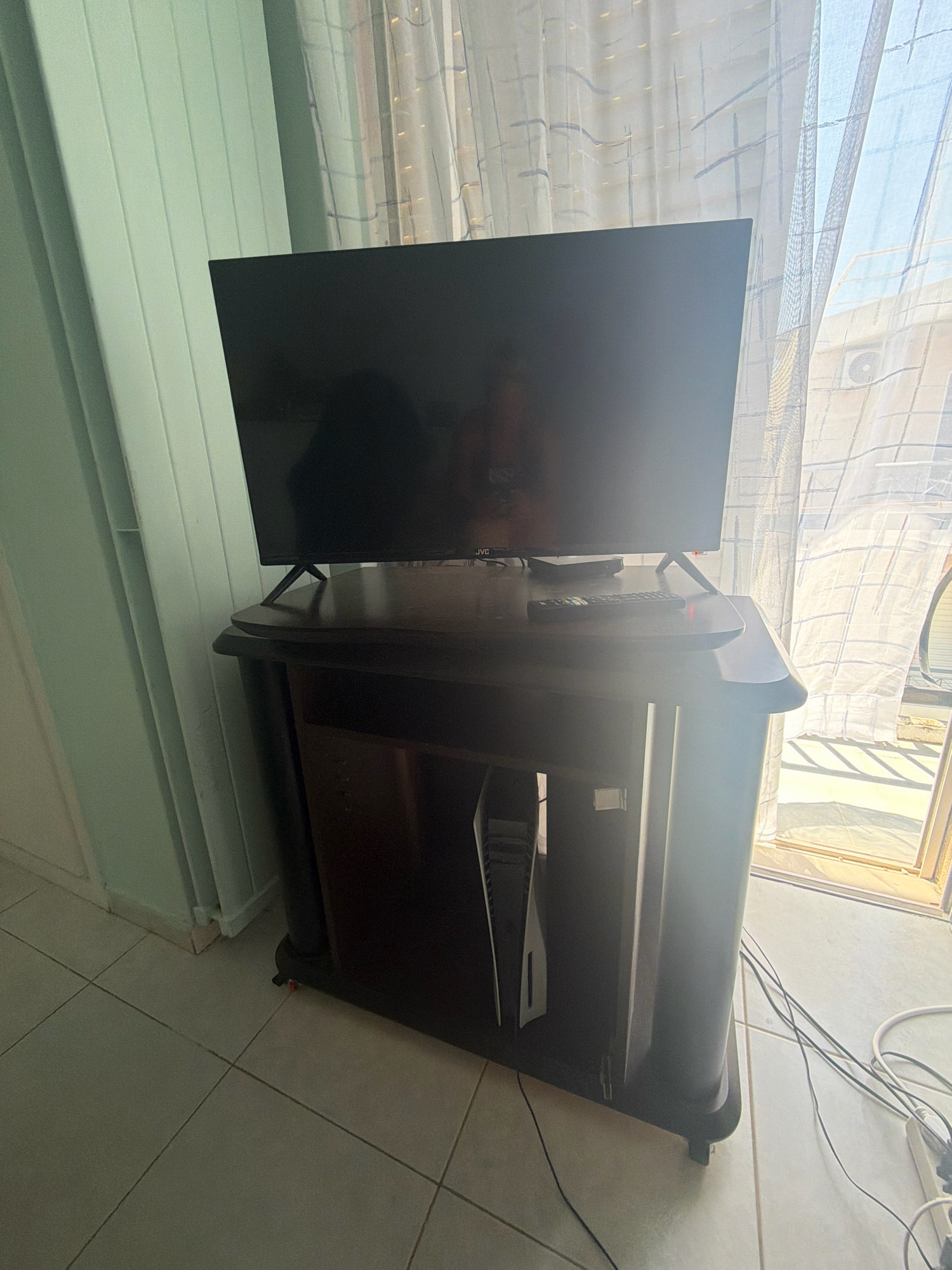 The image shows a television set on a dark wooden TV stand. The TV is black and has a JVC logo on the front. A remote control and a small black box are on the surface of the TV stand. To the left of the TV stand, there are light green vertical blinds. To the right of the TV stand, sheer curtains are drawn across a window, revealing a bright exterior with architectural elements and an air conditioning unit. Inside the TV stand, a white game console with a black controller is visible. The floor is tiled with light-colored square tiles. The overall scene appears to be an indoor living space, possibly in an apartment in Chalandri, Greece, given the context. There are no people present in the image.