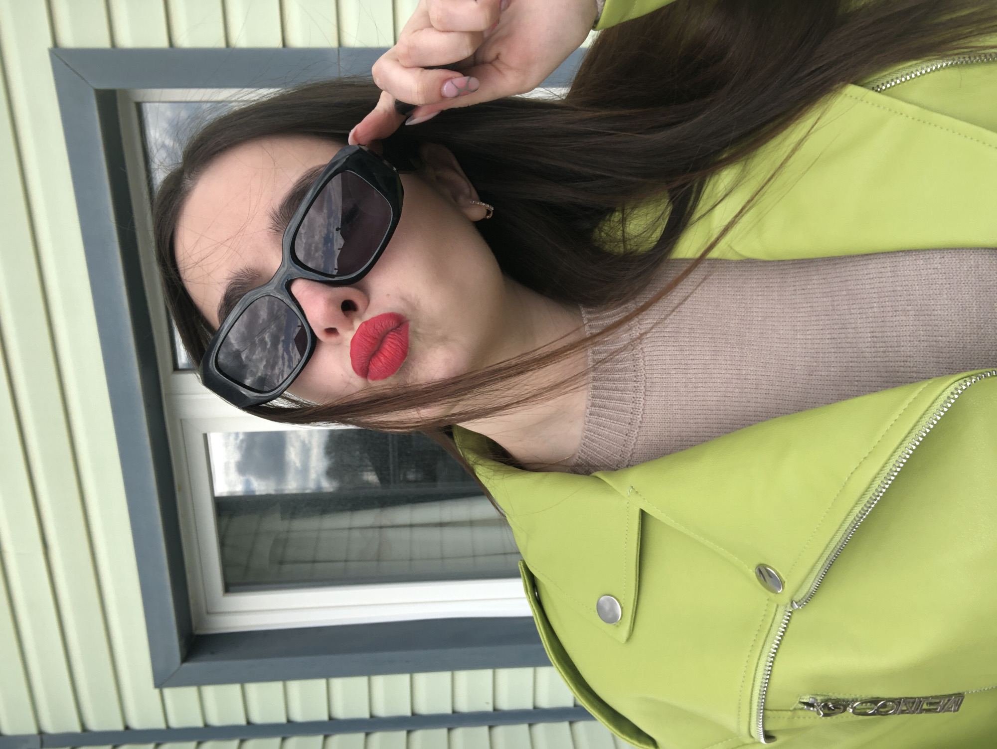 This vertical selfie features a person, likely female, captured from the chest up, posing outdoors in what appears to be Noviy, Russia. The individual has long, dark brown hair falling over their shoulder and is wearing large, black-framed sunglasses that reflect a cloudy sky. Their lips are distinctly painted with red lipstick and are pursed in a pout. One hand, with manicured nails featuring a light base and abstract dark lines, is raised near their temple, seemingly adjusting the sunglasses or hair. A small, simple hoop earring is visible in the ear.
The subject is dressed in a vibrant lime green (or neon green) moto-style jacket, detailed with silver zippers and snaps. A light brown or beige knitted sweater is visible underneath.
The background consists of the light, vertically-paneled siding of a building. A window with a dark grey outer frame and a white inner frame is visible behind and to the left of the subject, also reflecting the cloudy sky, indicating an overcast daytime setting. On a zipper pull of the jacket, the brand name "MENCOR" is visible. The pose and attire suggest a playful and stylish mood.