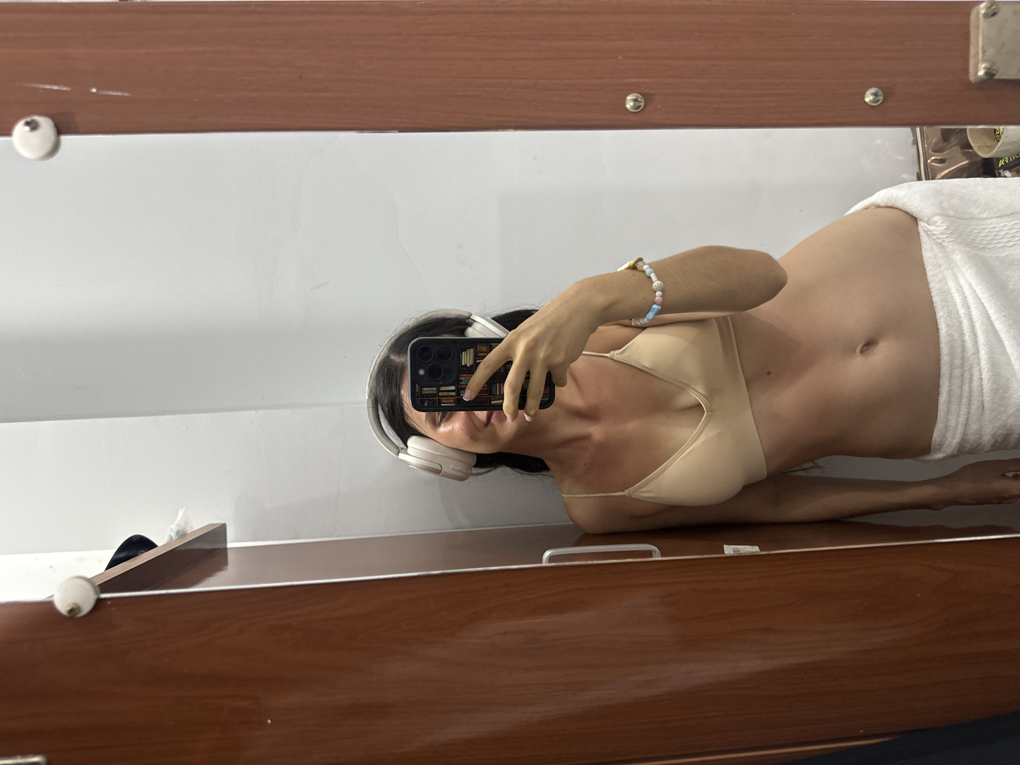 A woman is taking a selfie in a mirror. She is wearing a beige bra, a white towel, and white headphones. She is holding a smartphone with a case featuring an image of a bookshelf. She is wearing a beaded bracelet on her wrist. The background consists of a white wall and a brown wooden frame. The location is likely indoors but Mũi Né, Vietnam cannot be determined based on the image.