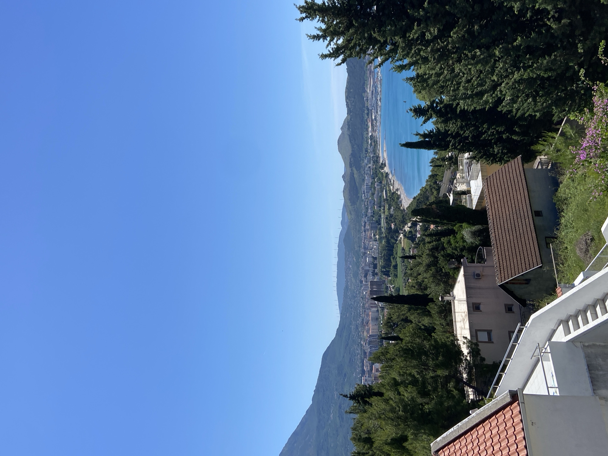View of Šušanj, Montenegro, with a clear blue sky above. The landscape includes a coastal area with a beach, a town with buildings visible along the coast, and a mountain range. The foreground shows parts of residential buildings with tiled roofs, white exteriors, and staircases. Trees, including coniferous species, are scattered throughout the scene. The sea is visible between the town and the horizon. No people are apparent in the image.