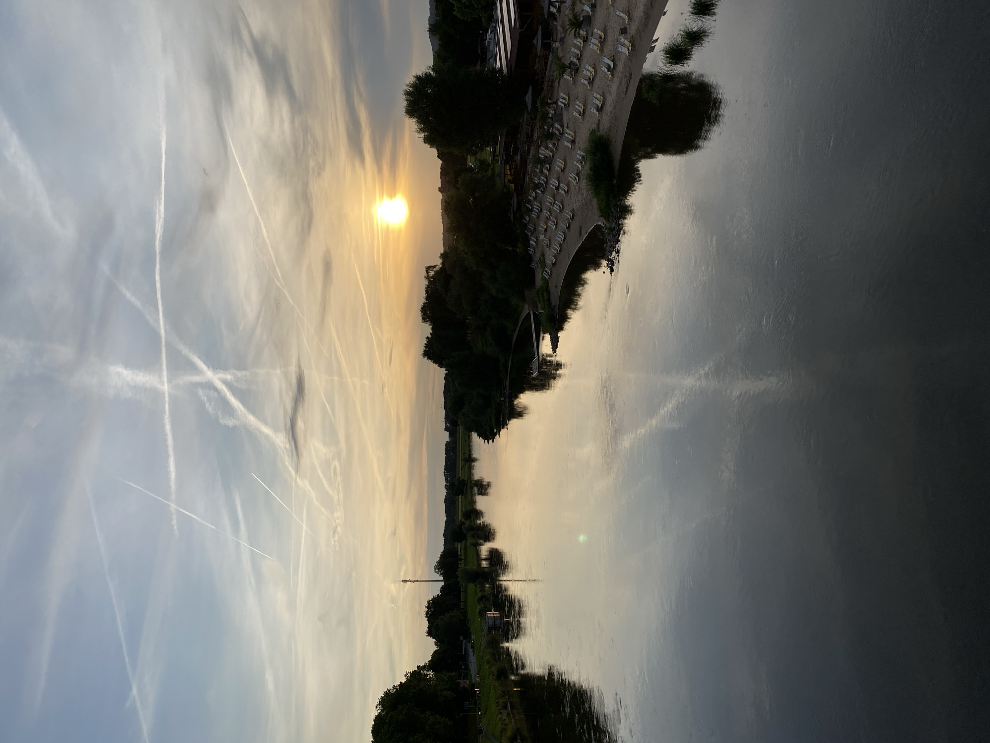 The image depicts a serene sunset over the Weser River in Rinteln, Germany. The sky is a canvas of soft blues and oranges, streaked with numerous contrails from aircraft, creating an intricate pattern. The sun, a bright orb, is low on the horizon, casting a warm glow on the scene.
On the right bank of the river, a sandy beach is lined with rows of empty lounge chairs, suggesting a beach club or recreational area. Trees and buildings form the backdrop of the beach. The left bank of the river features a line of trees and a distant telecommunications tower. The tranquil river water reflects the colors of the sky and the surrounding landscape, creating a mirror-like effect. The overall atmosphere is peaceful and picturesque.