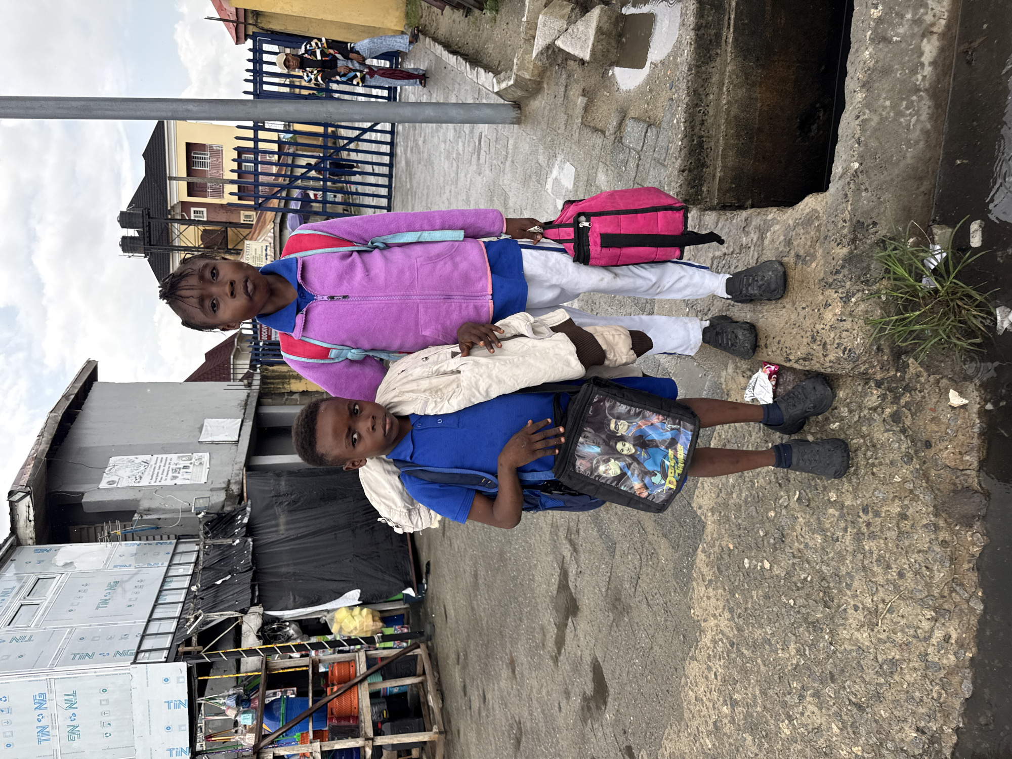 Two children stand side-by-side on a concrete slab next to a drainage ditch on a street in Ado, Nigeria. The child in the foreground wears a blue shirt, white jacket draped over the shoulder, and a black lunch bag with cartoon characters on it. The child in the background is wearing a purple jacket, blue shirt, white pants, and carrying a pink backpack. Buildings, including one with a corrugated sheet metal façade, line the street behind the children. In the background is also a blue gate.