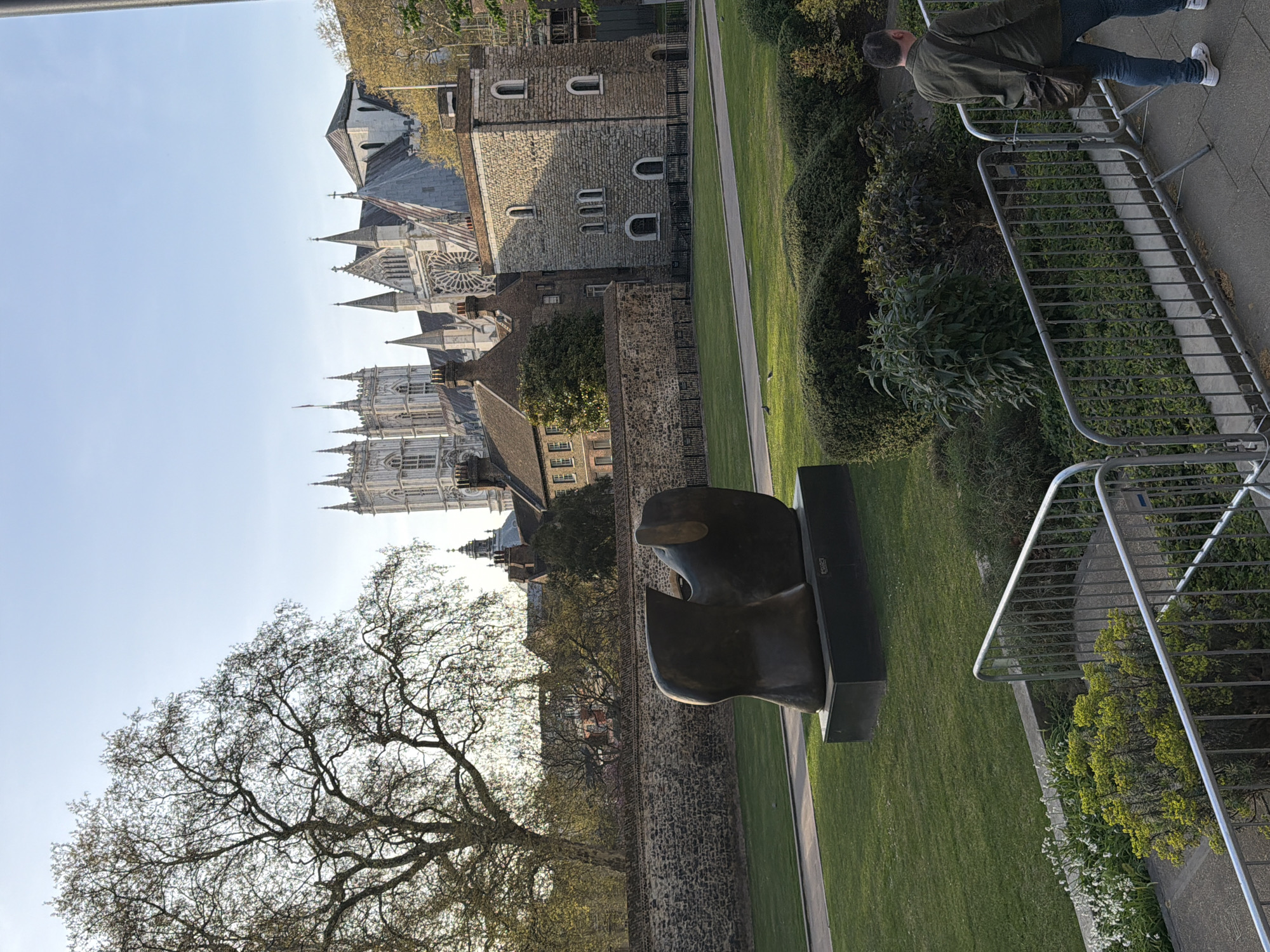 This outdoor shot captures a portion of London, United Kingdom. Westminster Abbey stands prominently in the background. The frame features a large, leafless tree and a modern sculpture in the foreground, set against a grassy lawn and a stone wall. There are metal barriers visible near the sculpture and the green area. A person is walking on the pavement in the upper right part of the frame.
