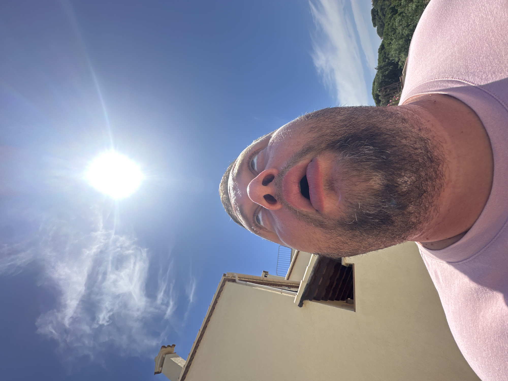 A man, with a full beard and open mouth, looks upwards at the bright sun against a clear blue sky. The sun is positioned in the upper center of the frame, emitting lens flares. To the left of the man's face is a section of a building with a beige facade and a terracotta-tiled roof. Further to the left, a chimney with a decorative top protrudes from the roof. In the background, lush green trees and foliage can be seen, suggesting an outdoor setting in a region with a Mediterranean climate. The man is wearing a pale pink t-shirt. The photograph was taken in Sant Feliu de Guíxols, Spain.