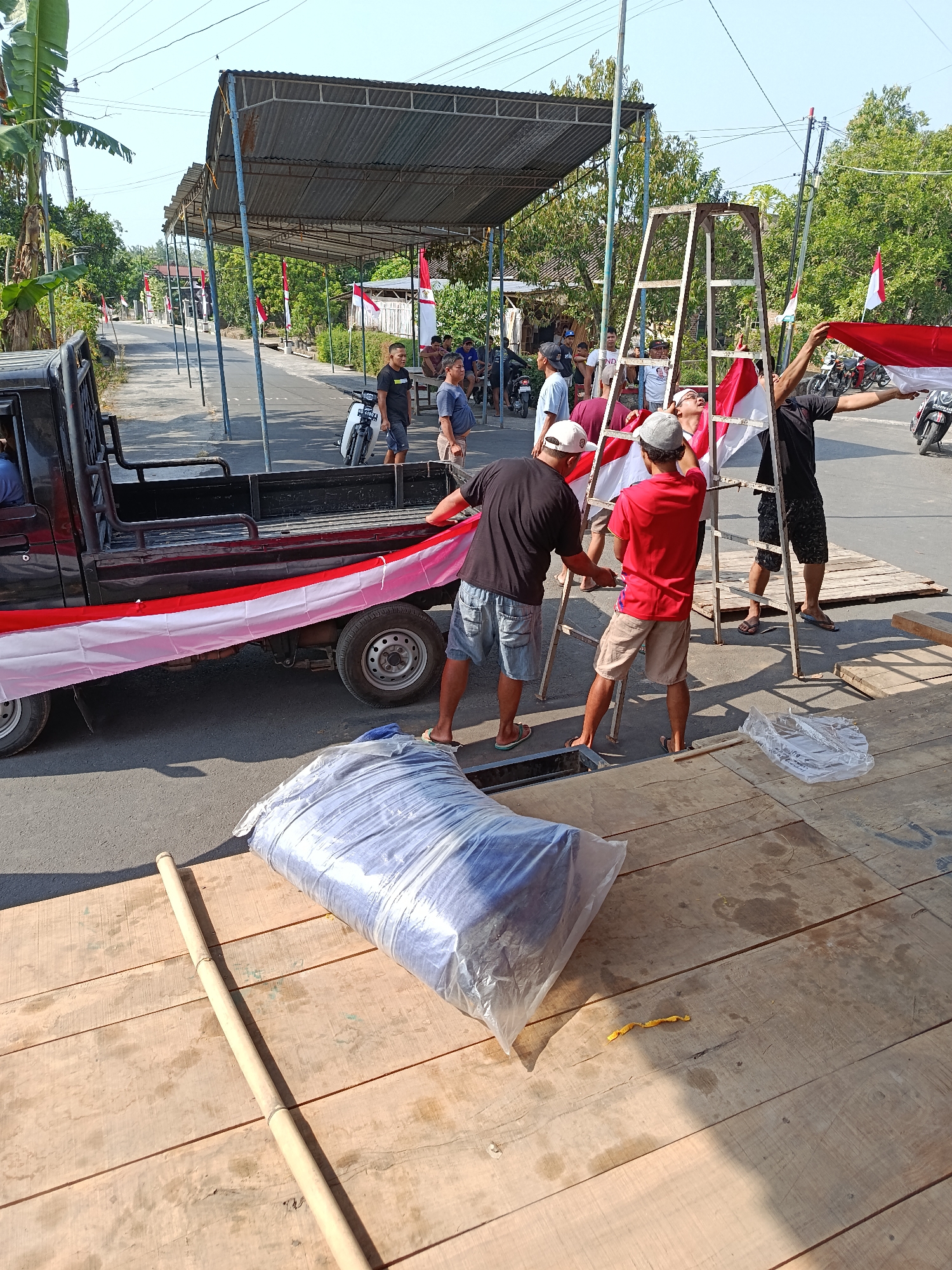 In the city of Jurug, Indonesia, several individuals are engaged in the process of raising an Indonesian flag. A red and white flag is being unfurled from a metal ladder, with multiple people assisting. The flag is also draped along the side of a red pickup truck, secured with white and red fabric. The scene appears to be taking place on a street, with a covered structure and trees lining the background. Wooden planks are laid out on the ground, and a large, plastic-wrapped item is also present. Several motorcycles are visible in the vicinity, and the sky is clear and blue.