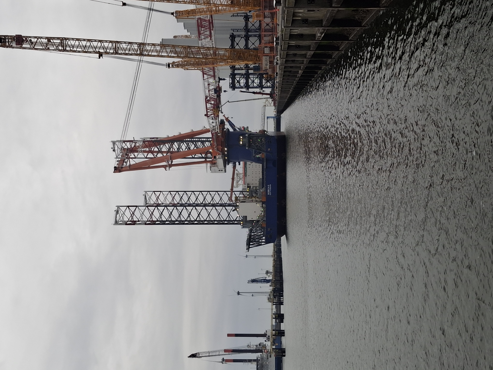 The image shows a harbor scene in Eemshaven, Netherlands, under a cloudy sky. The water surface is choppy. A large crane vessel, with a visible "BOREAS ROTTERDAM" inscription, is docked alongside a concrete pier. Various cranes, including orange and yellow ones, are visible in the background along the dock. Some wind turbine blades are also visible in the distance.