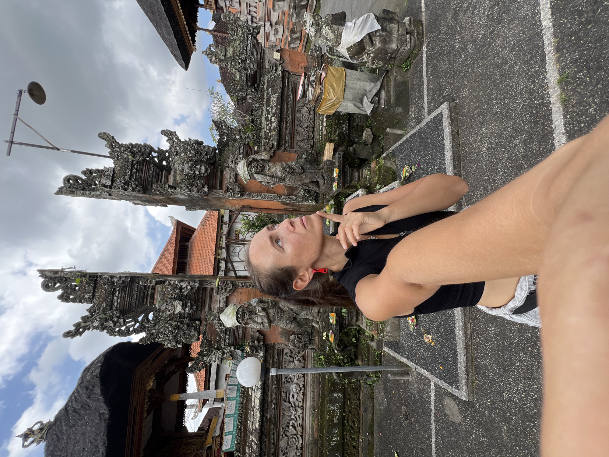 A sideways outdoor photo shows a light-skinned person with long brown hair in a black shirt in front of Balinese structures. The person is holding up their right arm and hand, appearing to take a selfie. Their face is turned to the right, and they are touching their chin with their finger. Behind them are two ornate stone structures with detailed carvings, typical of Balinese architecture, covered with moss and vegetation. The sky is overcast, and the ground appears to be made of asphalt. The location is likely in the Ubud District of Indonesia, given the distinctive style of the buildings.